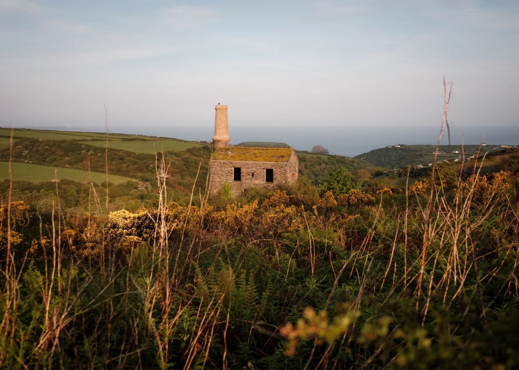 An abandoned stone Cornish mill stands among hills covered in heather. In the distance, you can just make out the hazy sea.