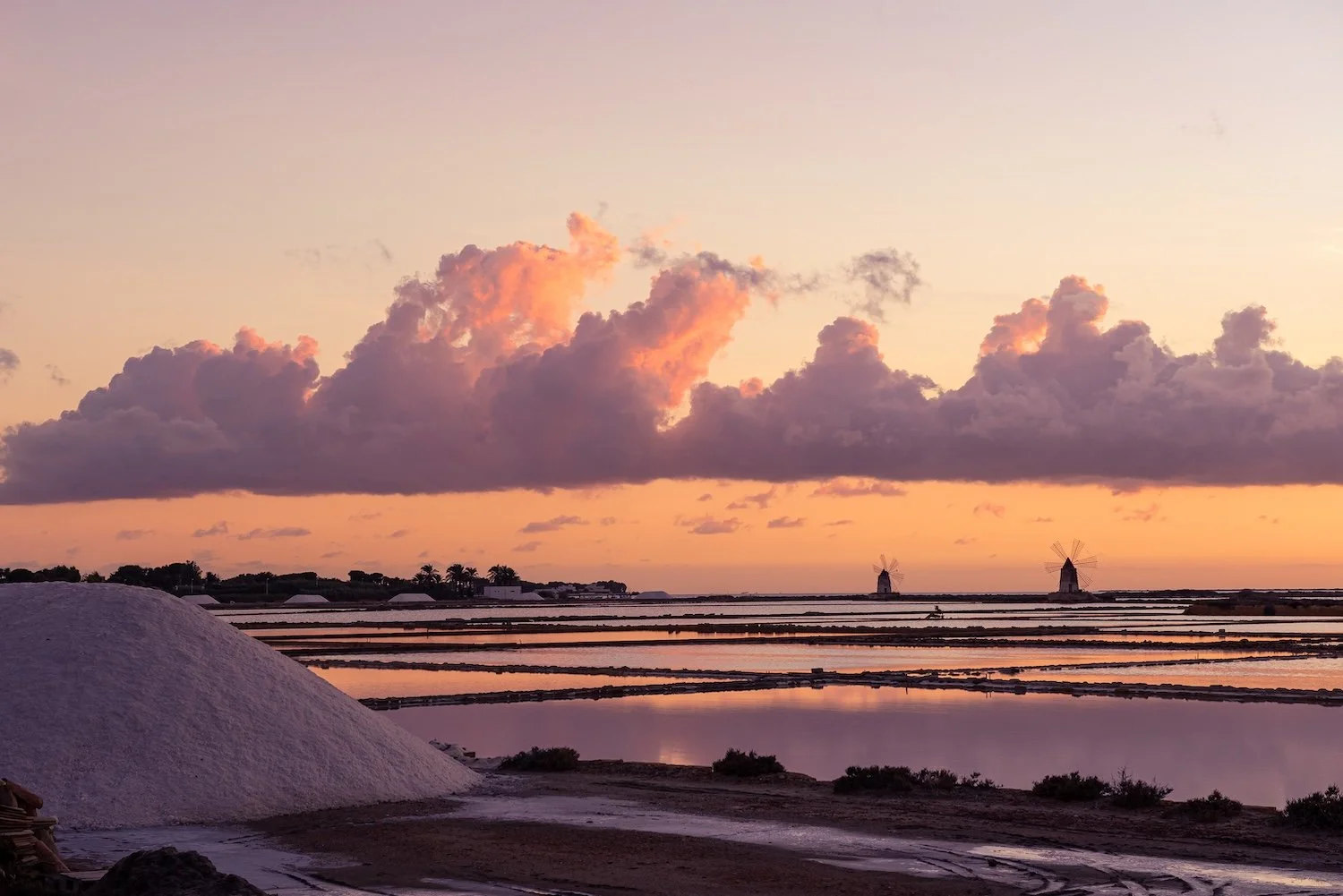 The salt plans of Trapani, Italy, reflect a pastel sunset, with the silhouette of windmills visible on the horizon