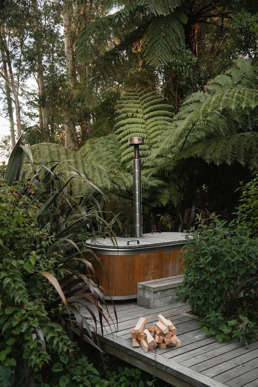 An outdoor hot tun is surrounded be green ferns. There is a pile of firewood in front of the tub to heat the water.