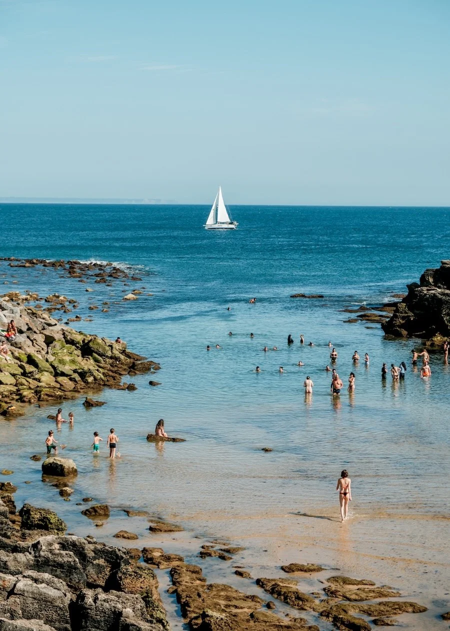 A white sail boat floats on the horizon as locals swim in a calm blue rocky cove