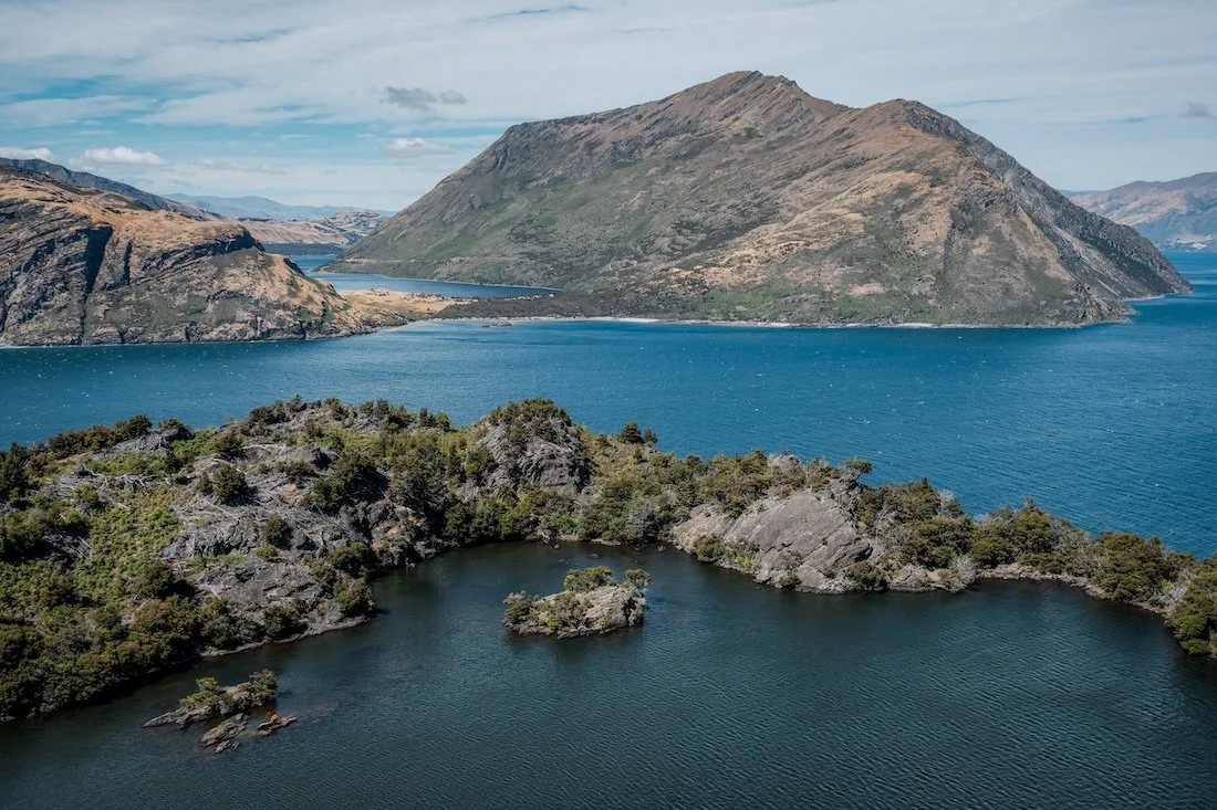 A series of tree-dotted rocky islands, broken up by bright blue water, photographed from Mou Waho Island in New Zealand's Wanaka