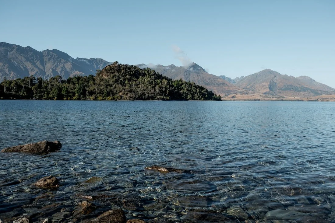 A crystal-clear lake, backdropped by green mountains, photographed in Queenstown, New Zealand