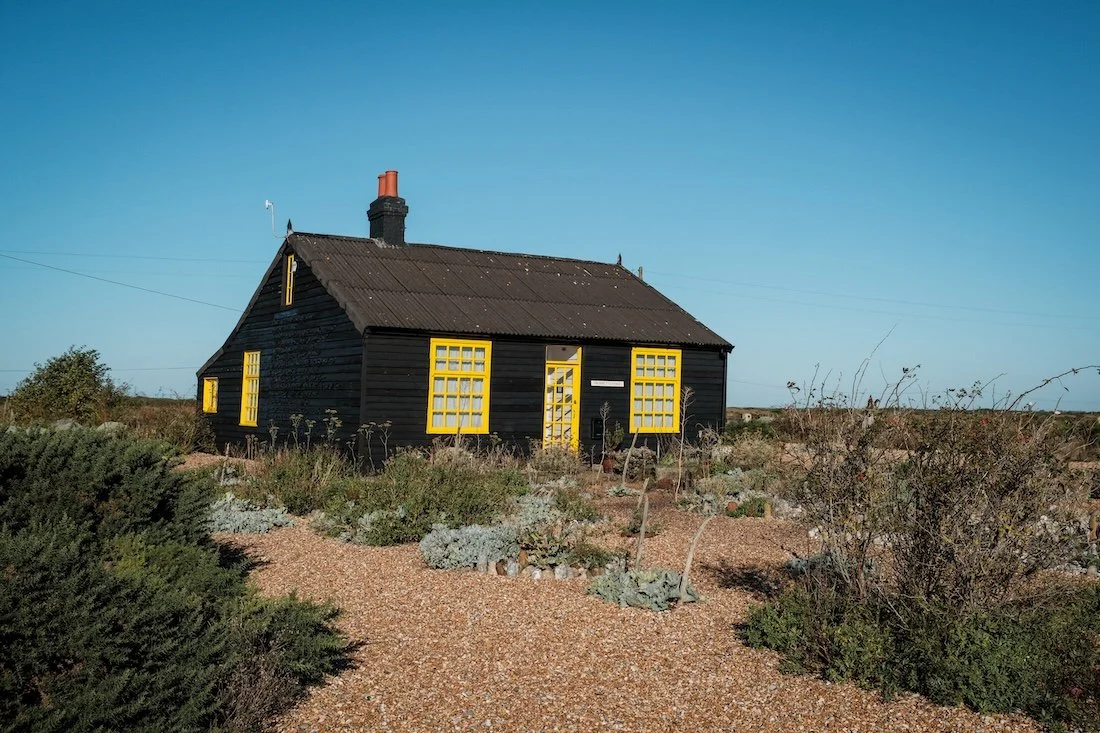 A black and yellow wooden cottage, once belonging to director Derek Jarman, stands among low green shrubs on a pebbly ground beneath a light blue sky