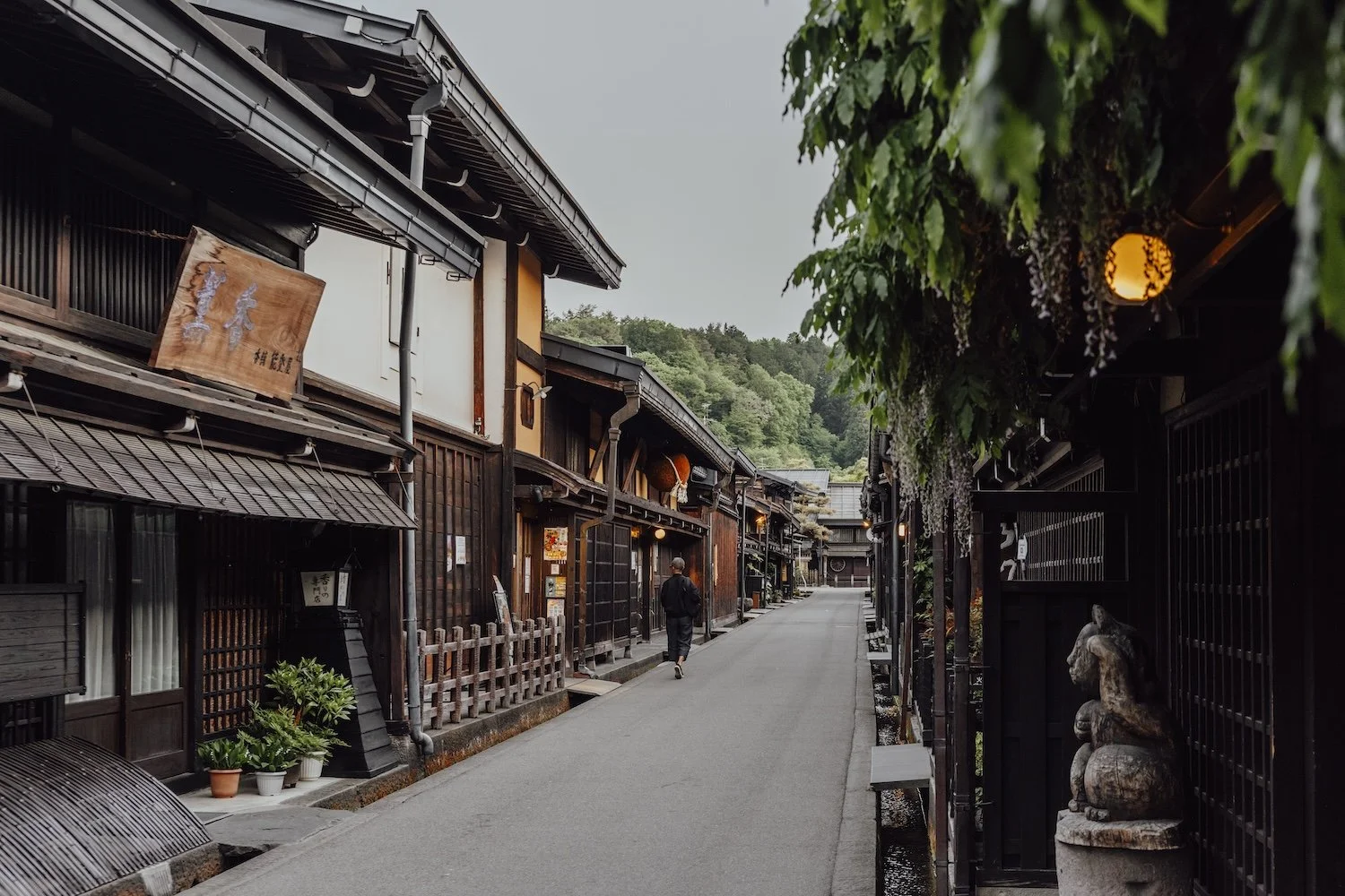 A lone traveller walks down a traditional Japanese street. Historic wooden buildings line and road and you can make out leafy mountains in the background
