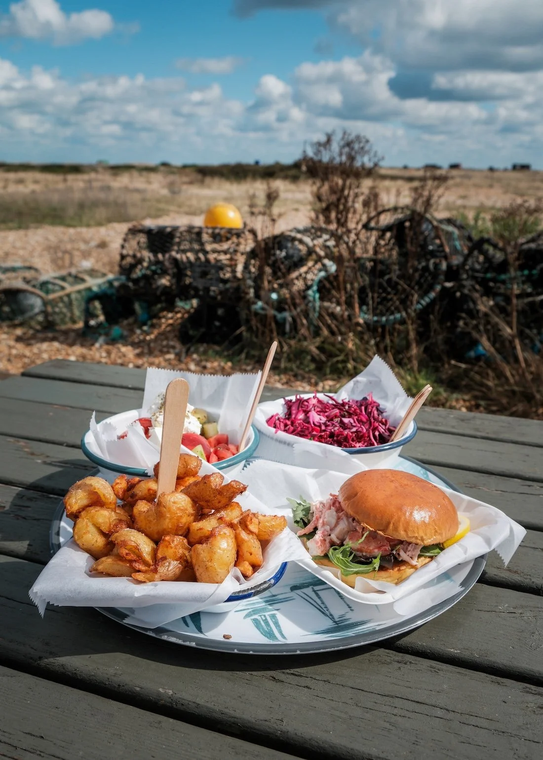 A fish burger, purple salad and crunch baked potatoes rest on a wooden table in the sun at pebbly Dungeness Beach