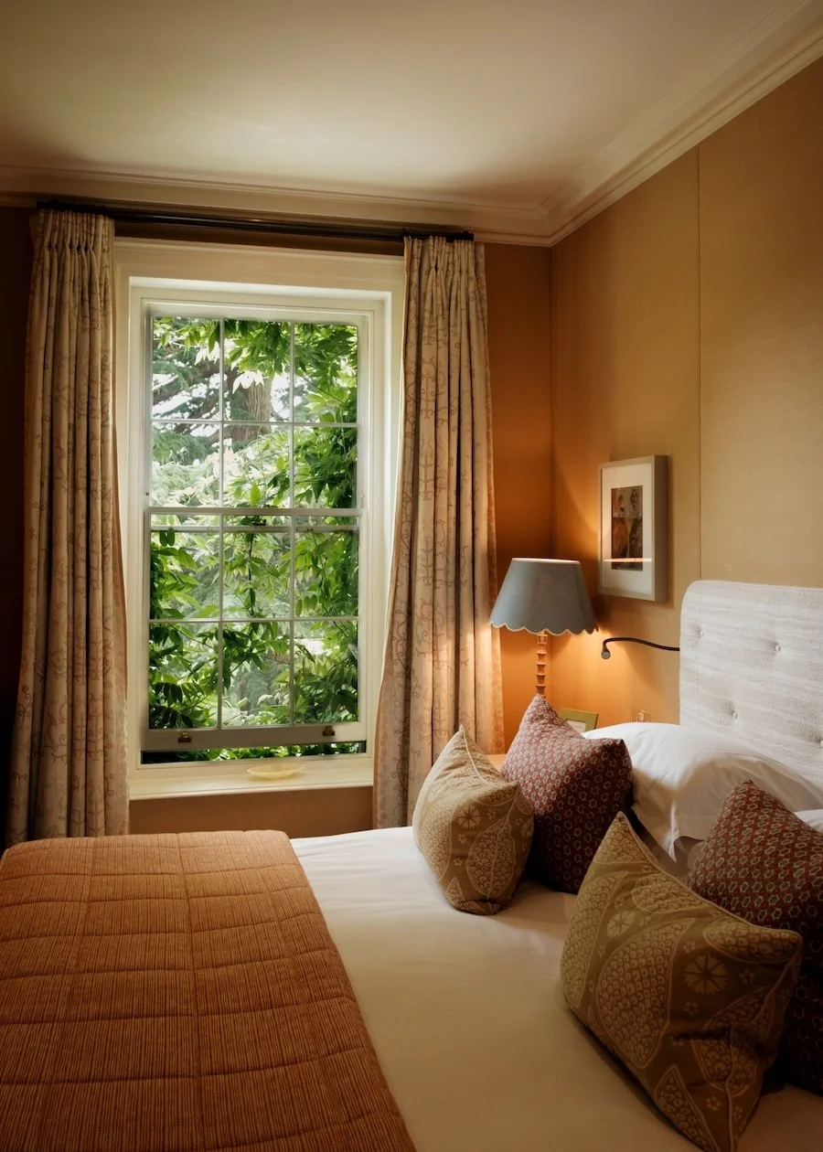 A close up of a bed, topped with orange and red pillows at Lime Wood in the New Forest. The window is slightly open and woodland can be seen through it