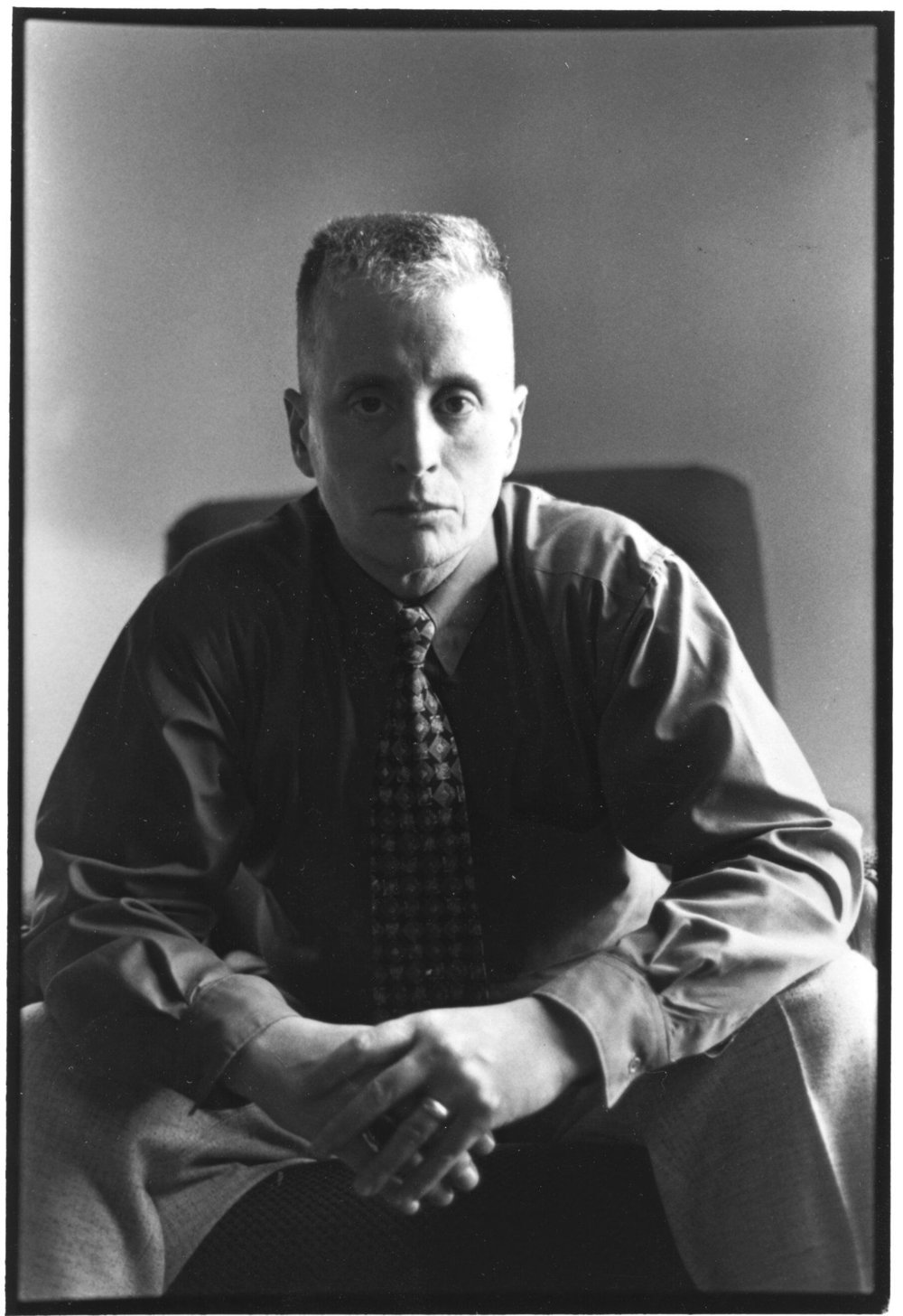 A black and white image of Leslie Feinberg sitting in a chair leaning forward slightly. She has a buzzcut and is wearing a button up shirt with a patterned tie, tucked into slacks. Her face is serious and she is frowning a little.