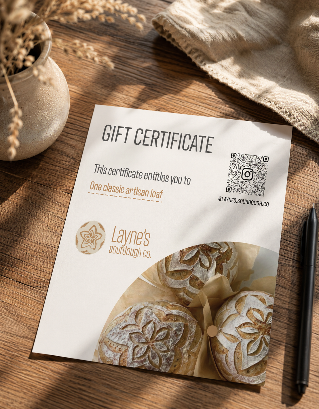 Gift certificate from Layne's sourdough co. for one classic artisan loaf, placed on a wooden table with a black pen, a beige cloth, a flower arrangement, and a photo of artisanal bread with intricate designs.
