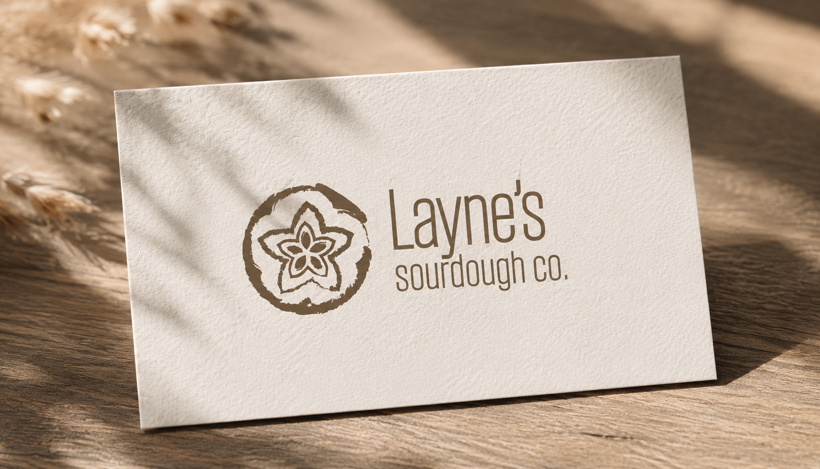 Business card for Layne's Sourdough Co. with a circular logo featuring a stylized flower. Card placed on wooden surface with natural lighting casting shadows.