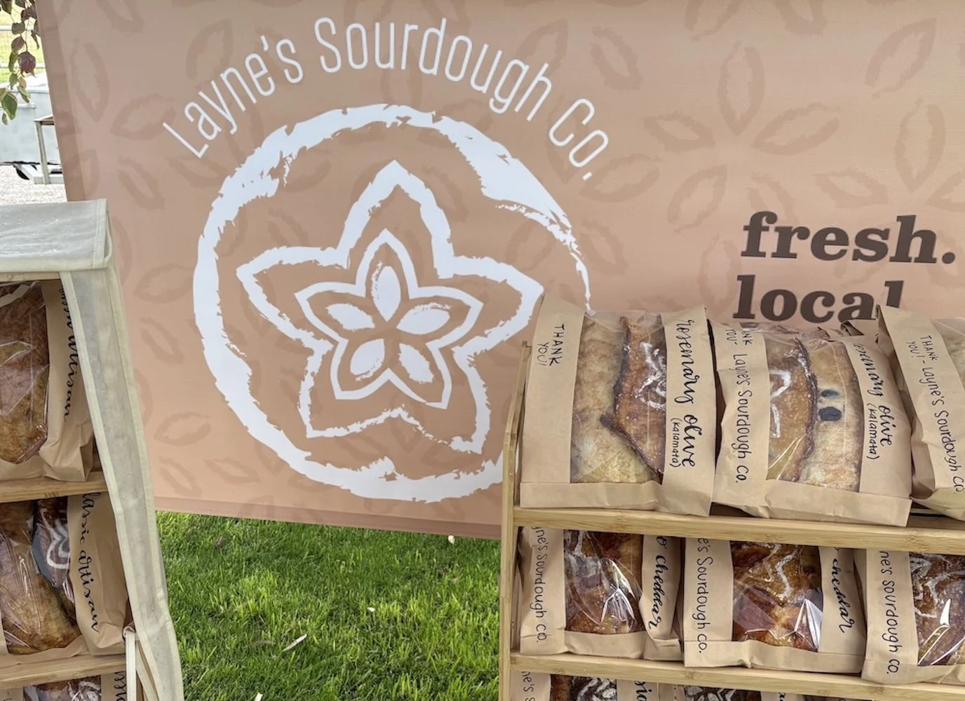 Display of baked goods labeled in paper bags on wooden shelves at Layne's Sourdough Co., with a large pinkish-beige banner in the background featuring the company logo and the words "fresh. local."