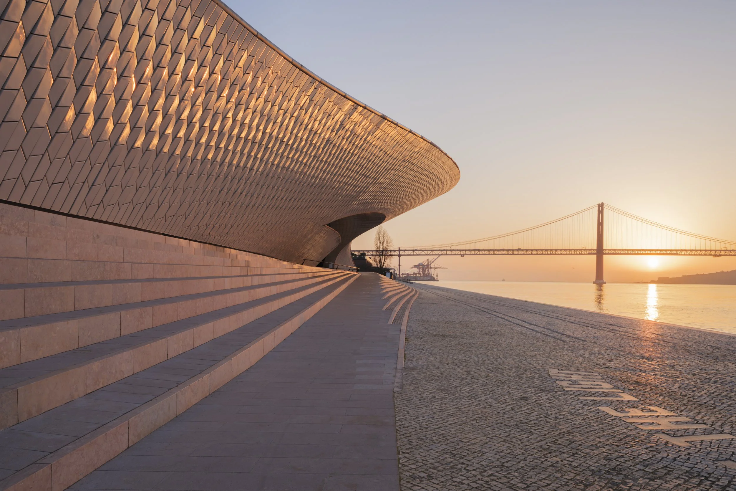 The first rays of sun light up the facade of MAAT with a golden glow. In the distance you can see the 25 de Abril Bridge spanning the Tagus river