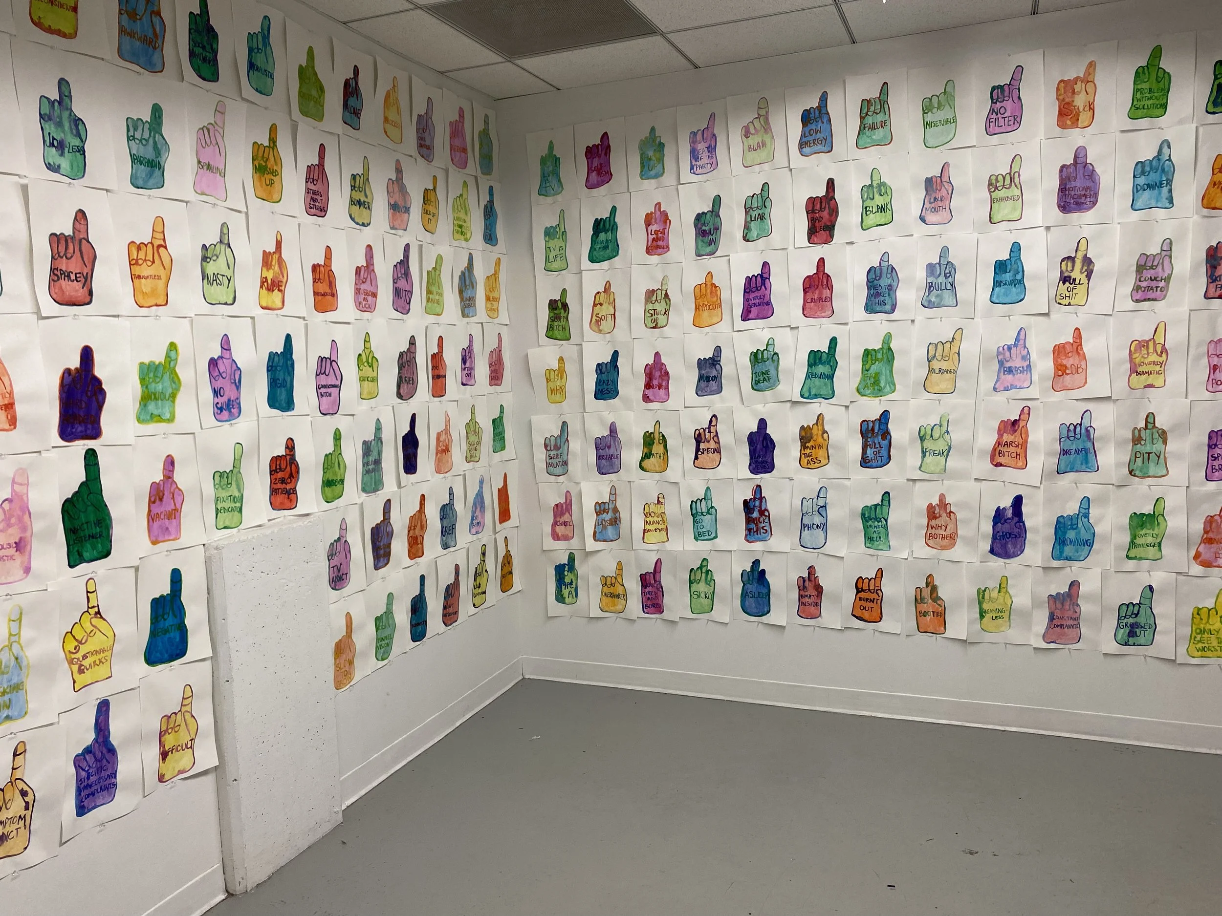   A detail shot of the corner of a white room with grey floors. The walls are wall papered with a grid of bright and colorful watercolor drawings on nine inch by twelve inch white paper. The drawings continue beyond the image making it seem like they