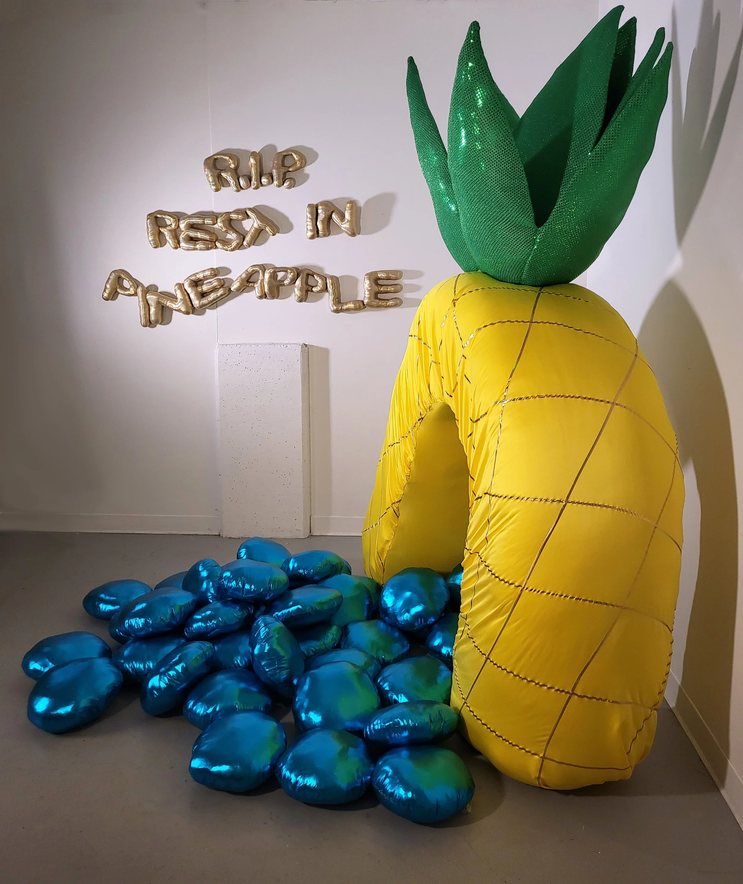   A profile view of a giant pineapple arch with sparkly green leaves and gold cross hatching on its yellow base stands on a grey floor. Spilling out from the arch are oversized turquoise spandex aquarium pebbles. On a white wall next to the pineapple