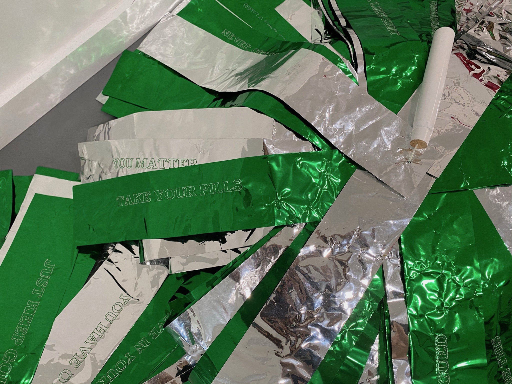   A close up image of giant pom poms with machine embroidered self talk phrases on them. Visible phrases include “YOU MATTER” and “TAKE YOUR PILLS.” The pom poms have silver and metallic green strands.  