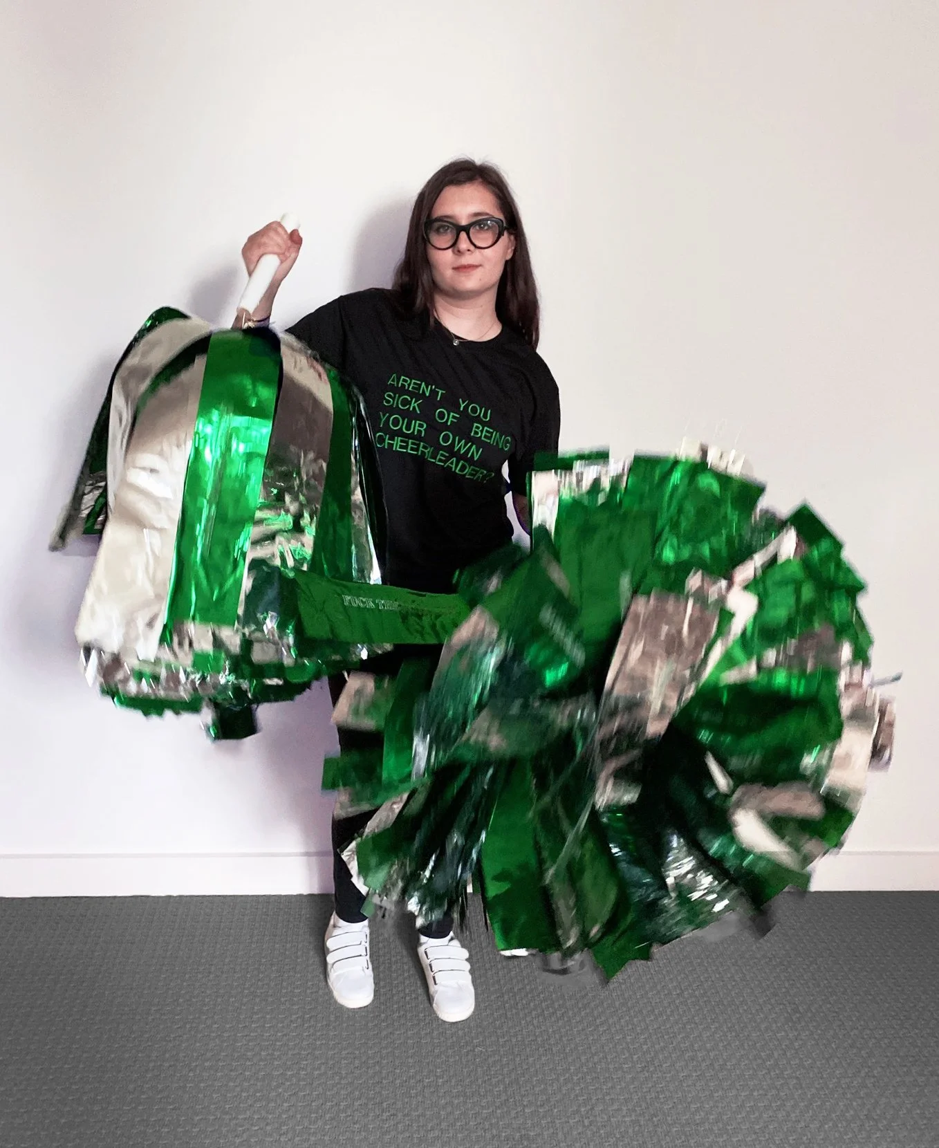   The artist, a white woman with brown hair and thick cat eye glasses, is wearing a black t-shirt with green embroidery on it that reads, “AREN’T YOU SICK OF BEING YOUR OWN CHEERLEADER?” In each hand, she clutches a giant silver and green mylar pom p