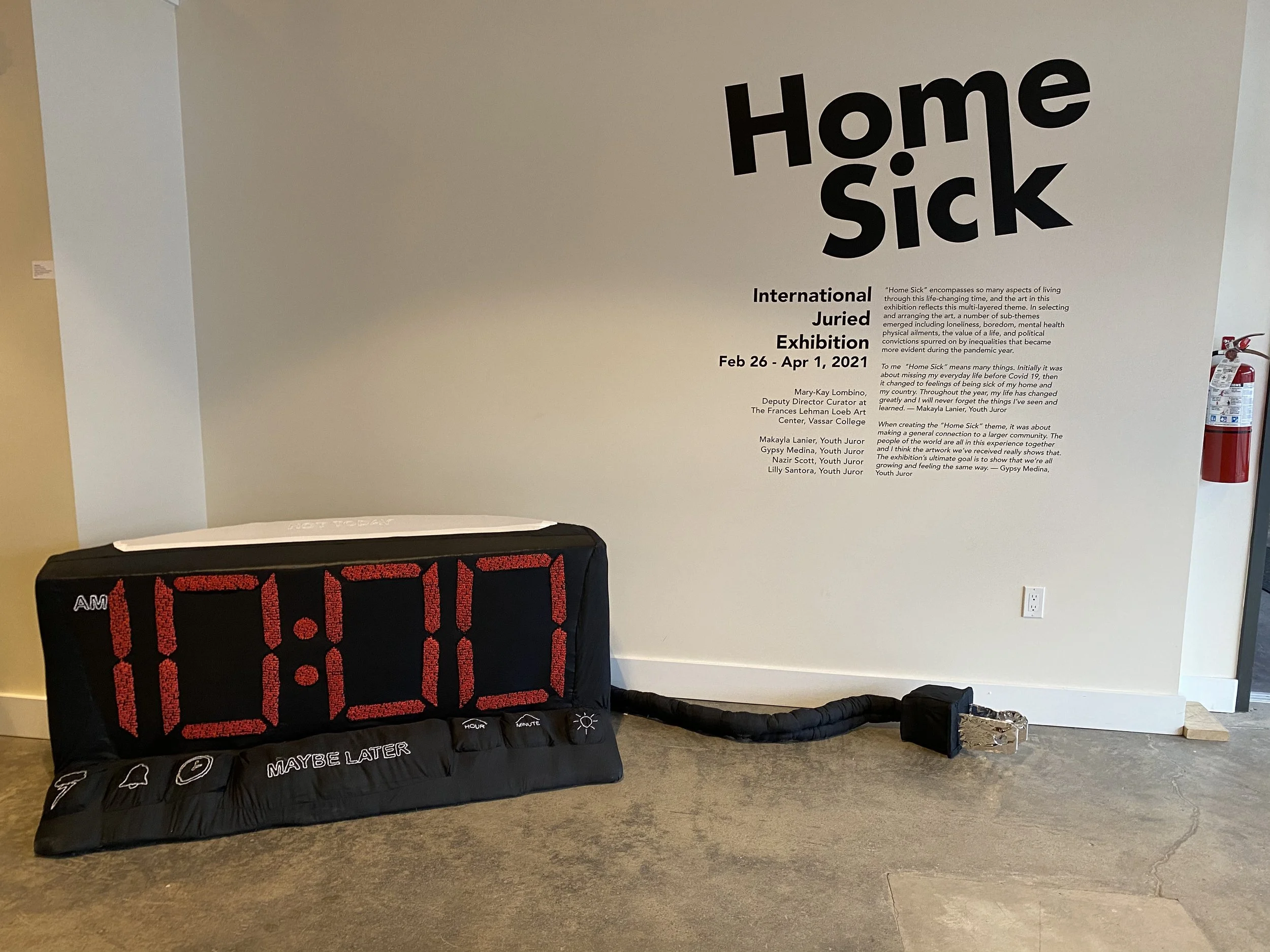   A white wall has exhibition text announcing the name of the show, “Home Sick” and other details about the exhibition such as dates and times, and other wall text that is too small to read. In front of the wall is a large soft sculpture digital cloc
