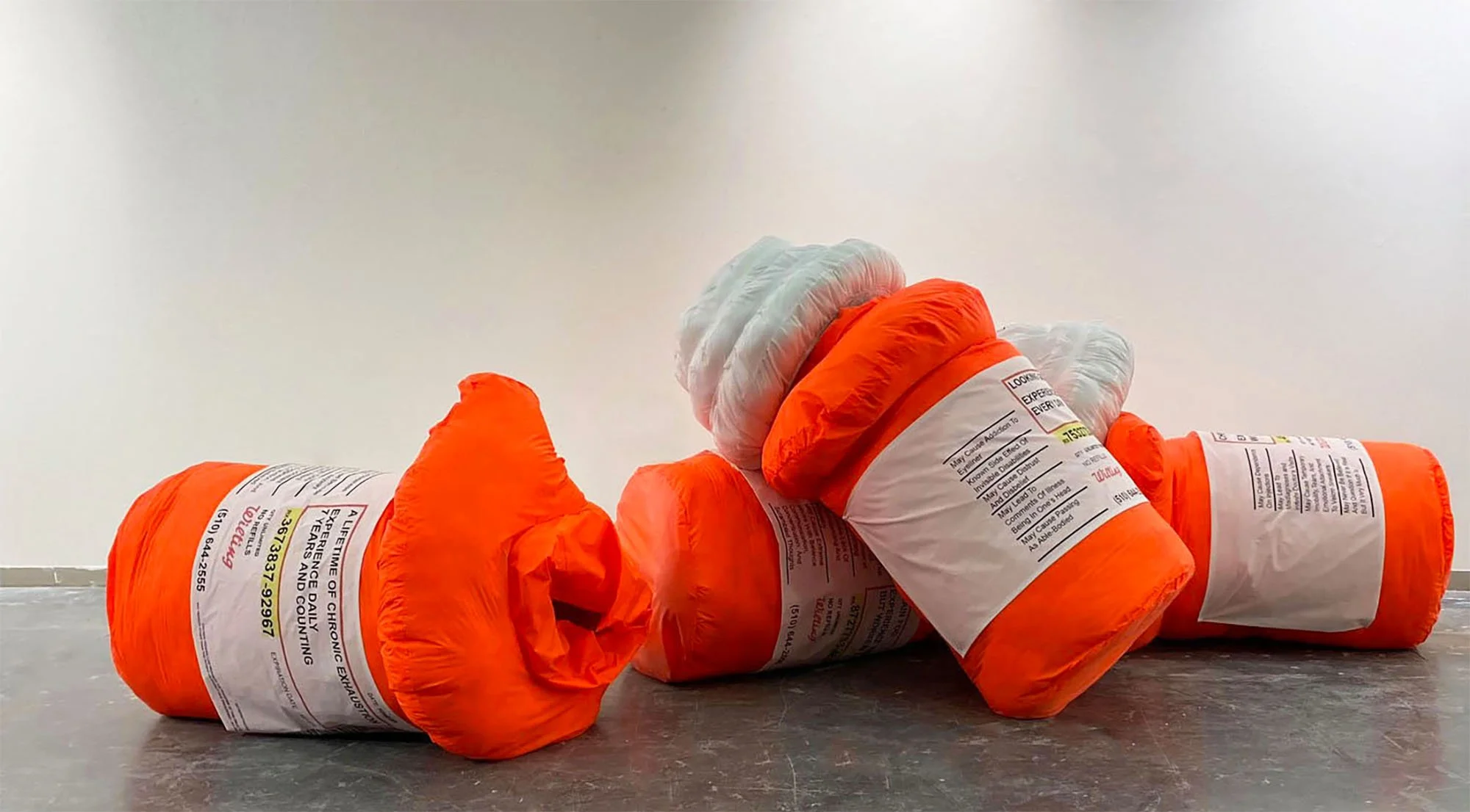   Four fluorescent neon orange nylon soft sculpture pill bottles are collapsed in a pill on the floor. One pill bottle is missing its cap revealing its hollow interior. The puffy pill bottles have custom labels that each represents a symptom of the a