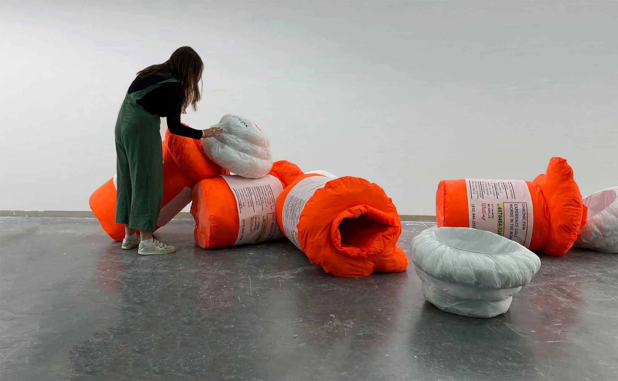   Four fluorescent neon orange nylon soft sculpture pill bottles are collapsed on a grey concrete floor. The artist a white woman with brown hair, leans over the bottles adjusting one of the caps. Each bottle represents a symptom of the artist’s chro