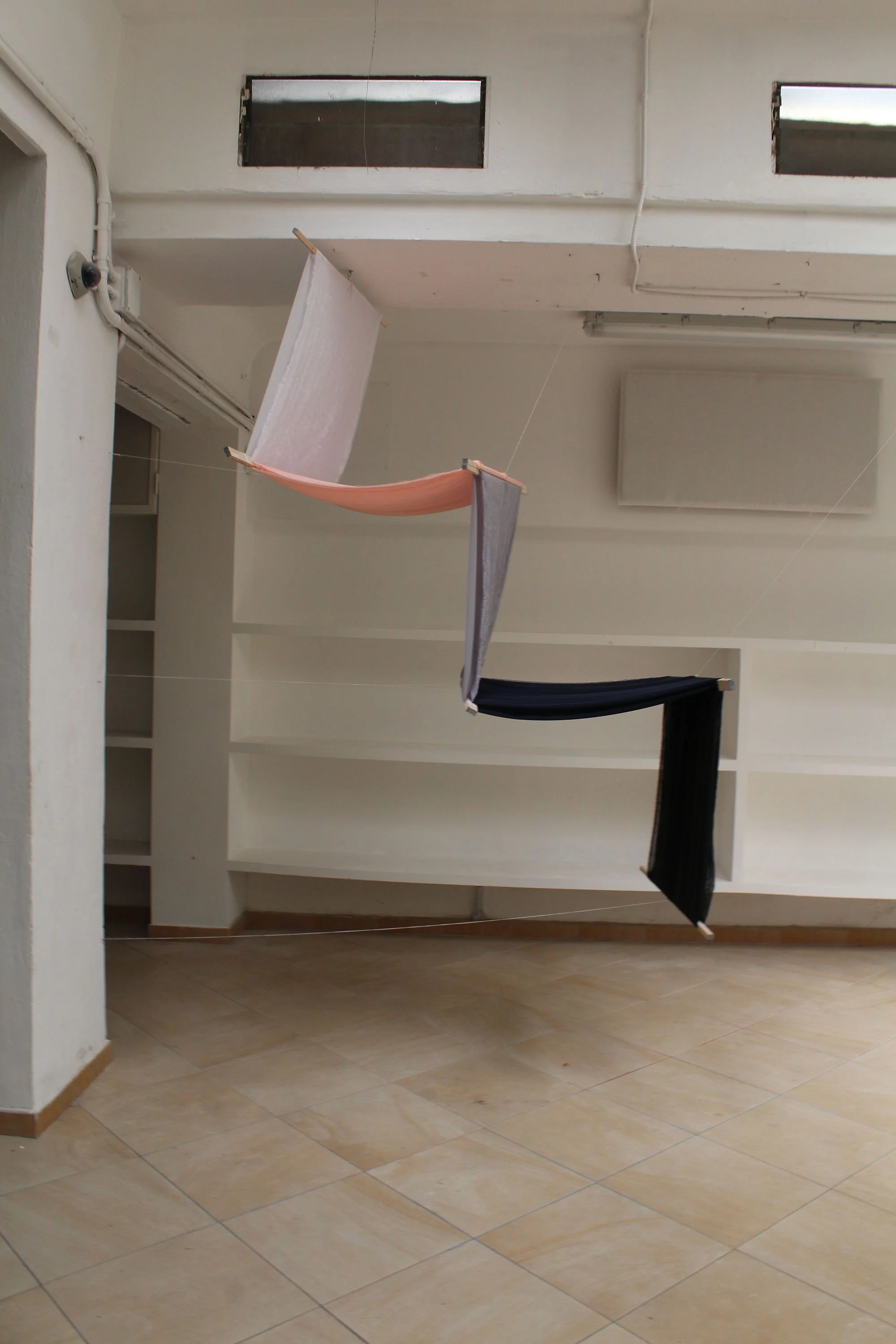   A white room with shelves on the wall has a sculpture suspended from its low ceiling. This photo shows the sculpture in a profile view. The sculpture is suspended in a way that mimics the shape of stairs and has white, pink, grey, navy and black ch