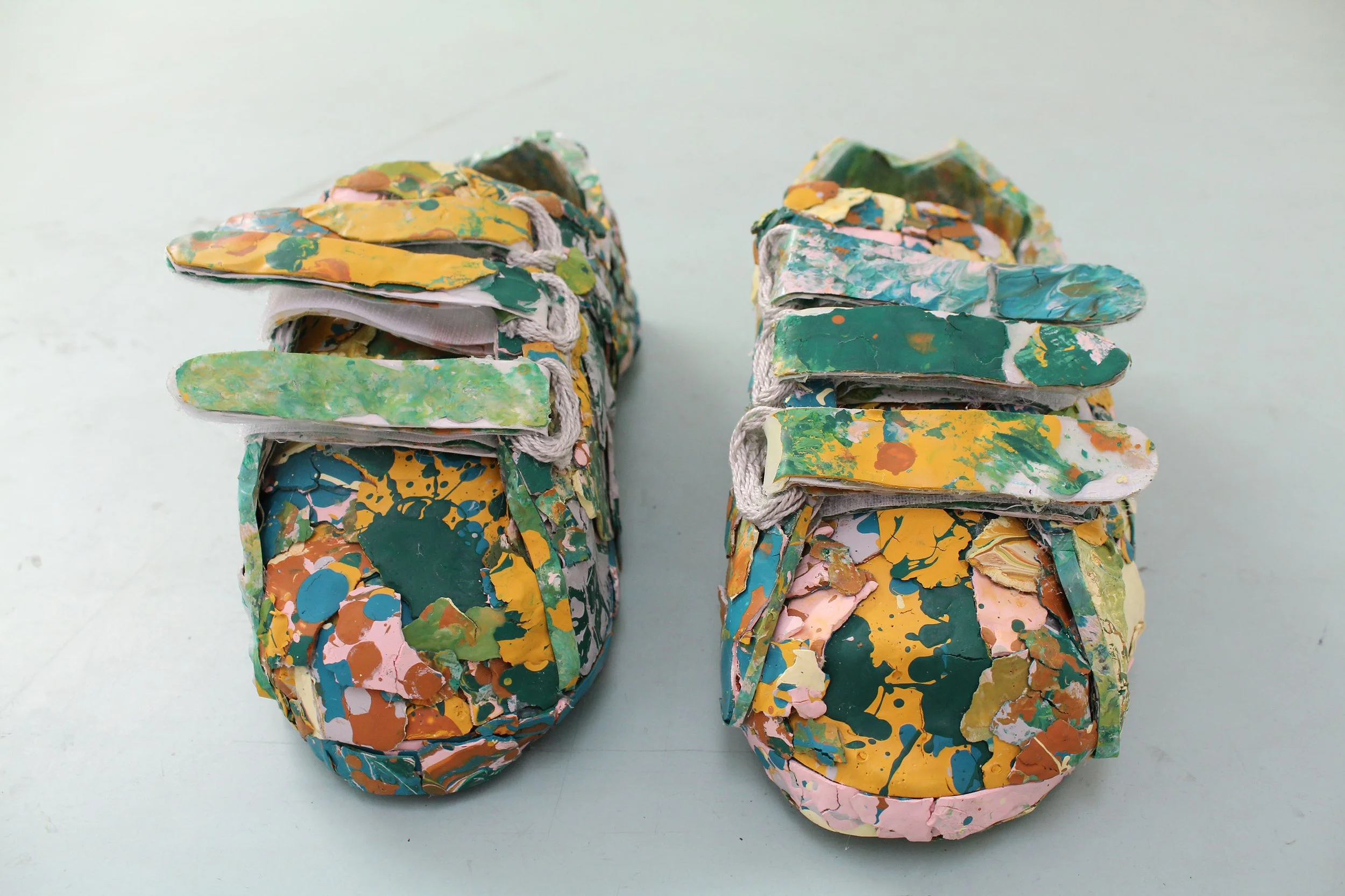   On a white background sits two velcro sneakers made of dried paint chips that give the visual impression of shows completely covered in paint splatters.  