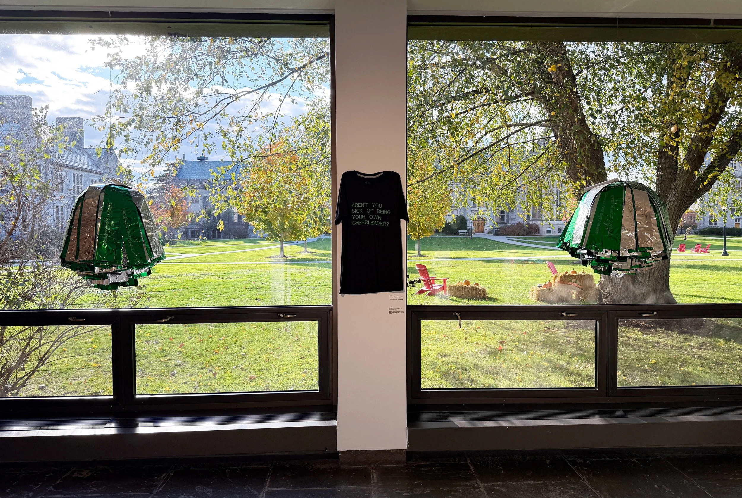  Hanging on a white post between two large windows is a black t-shirt. In kelly green embroidery, the t-shirt reads “AREN’T YOU SICK OF BEING YOUR OWN CHEEALEADER?”. Hanging in front of each window is a giant silver and green pom pom. 
