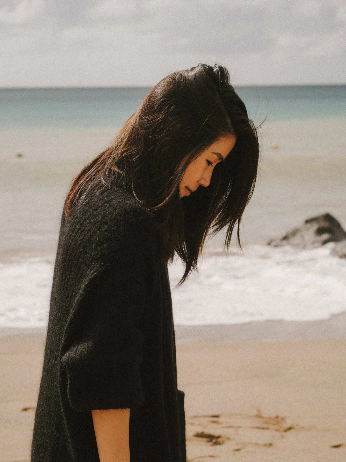 A woman with long dark hair standing on a beach, looking down, wearing a black sweater, with the ocean and sky in the background.