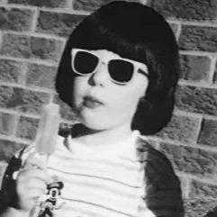 A person with short black hair, wearing white sunglasses and a striped shirt, making a peace sign with their hand in front of a brick wall.