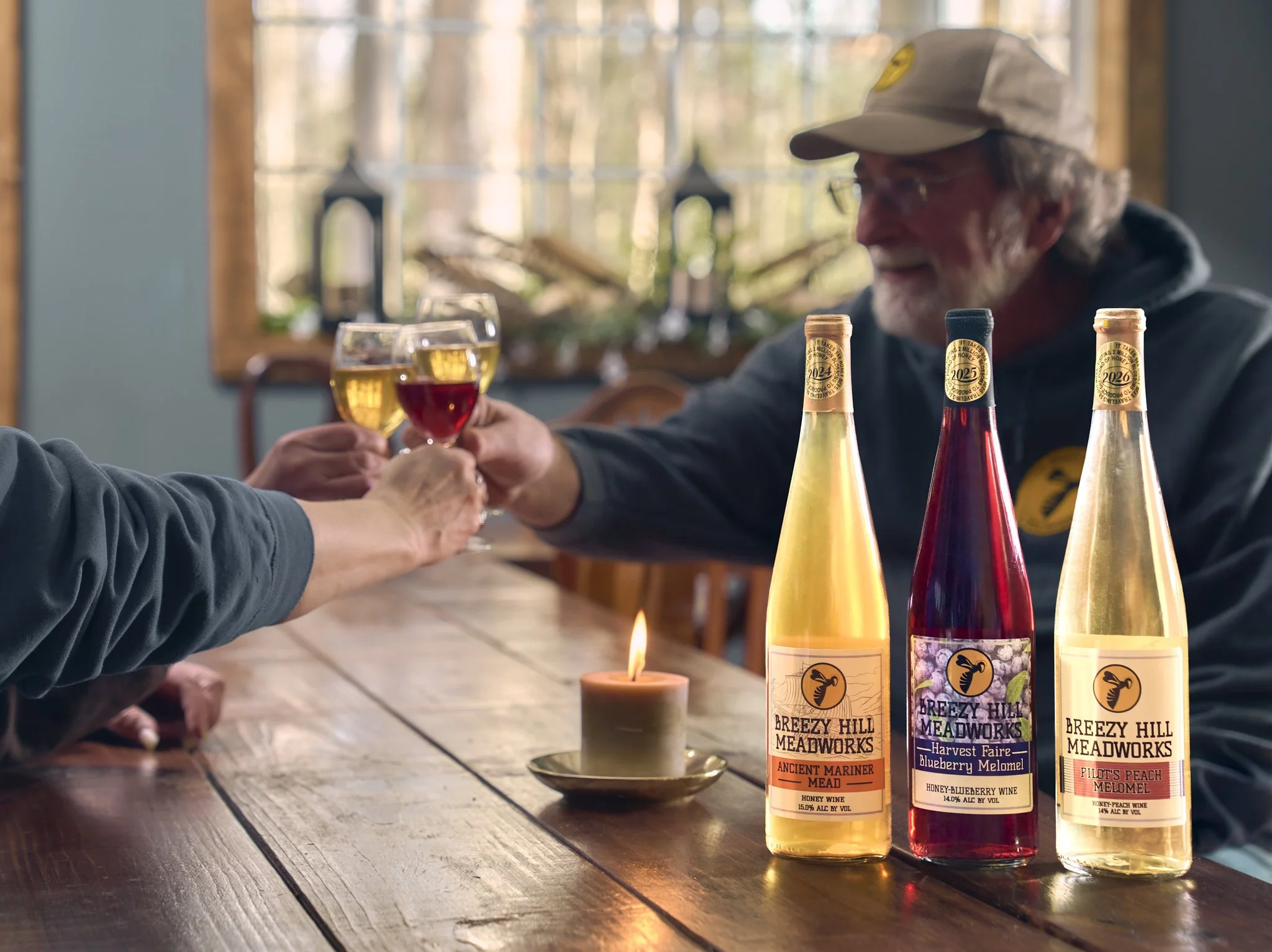 Two people clinking glasses of honey wine in a cozy setting with three bottles of Breezy Hill Meadworks on the table, a small lit candle, and a window showing outdoors.