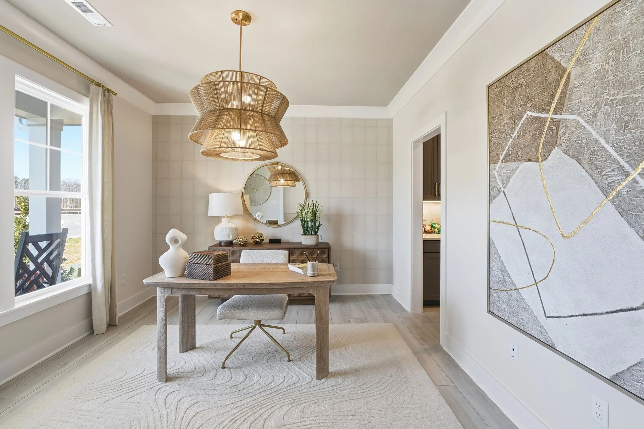 Modern office room with a wooden table, gray chair, large beige woven pendant light, white curtain on window, abstract art on wall, round mirror, table lamp, vase with greenery, and minimalist decor.