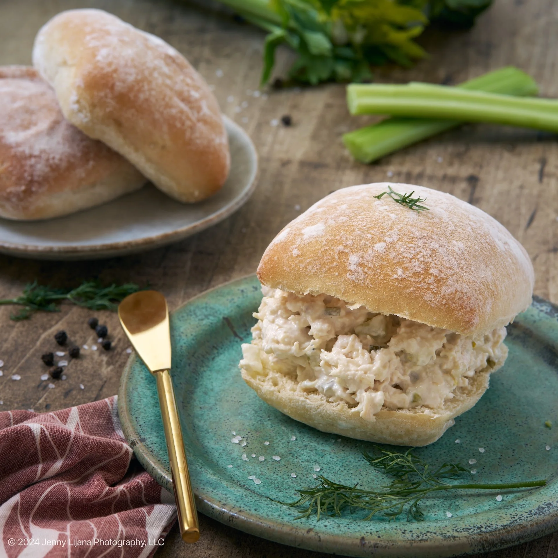 Chicken salad sandwich on a green plate with a small sprig of rosemary, a gold spoon, salt, and black peppercorns, with bread rolls on a plate in the background, on a rustic wooden table.