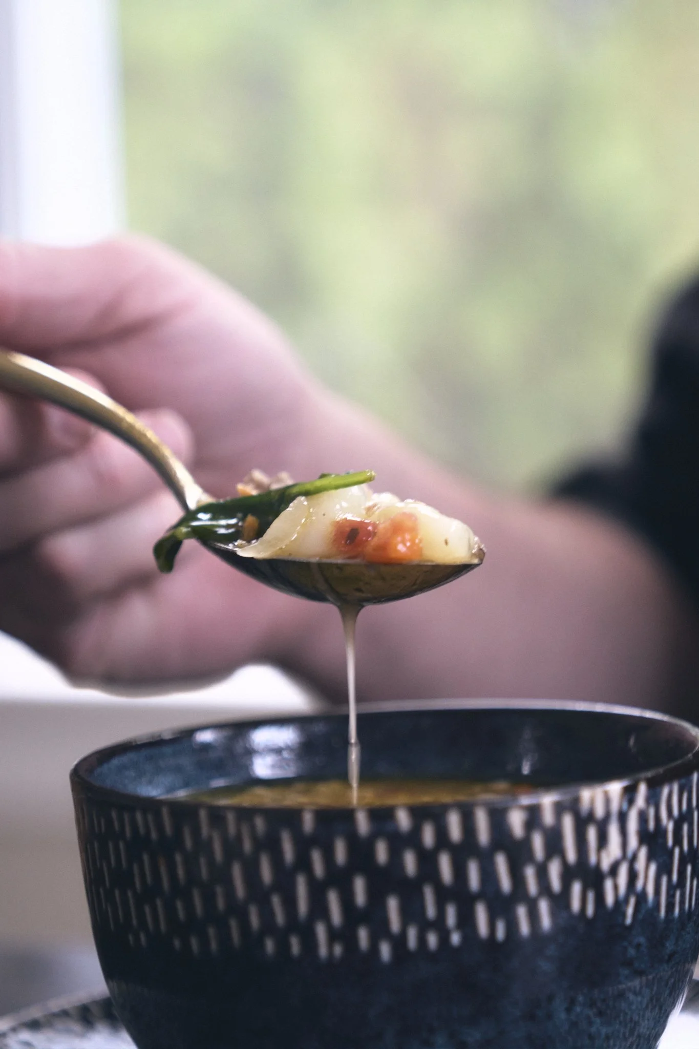 A hand holding a spoon with food above a dark blue bowl, with a stream of soup dripping from the spoon into the bowl.
