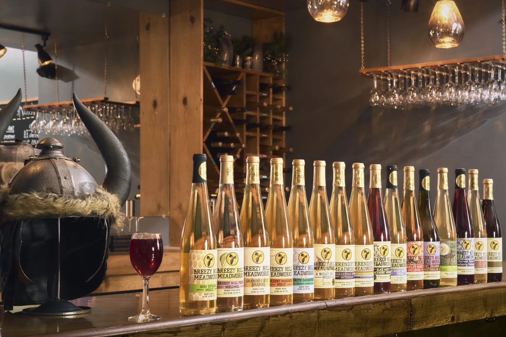 Row of Breezy Hill Meadworks bottles with different labels, a glass of red beverage, a Viking helmet with horns, and wine glasses hanging upside down from a rack in a cozy bar or restaurant.