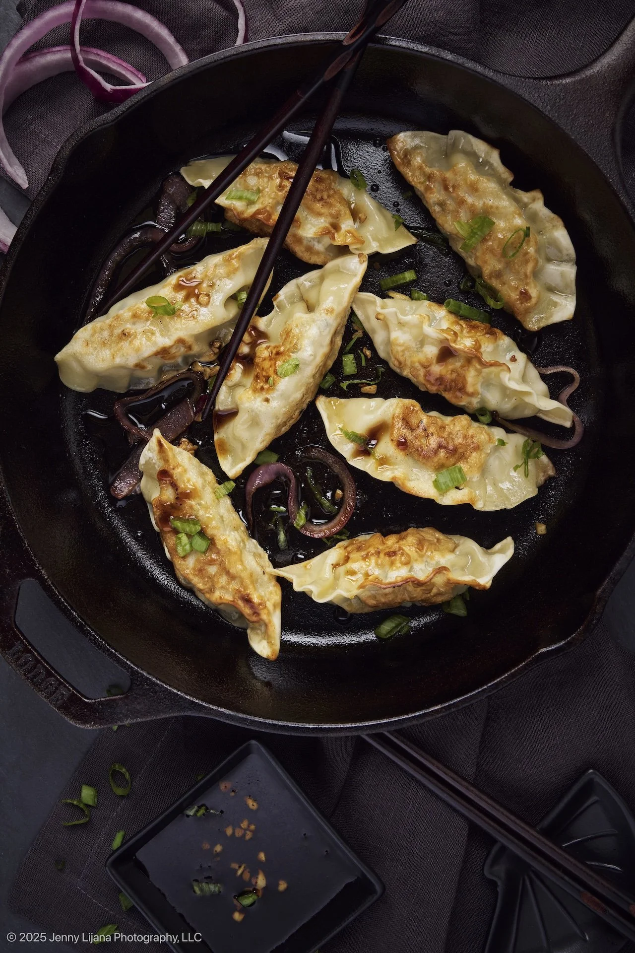 Pan of fried dumplings garnished with chopped green onions in a black cast iron skillet, with soy sauce and chopped garlic on a black square plate nearby.
