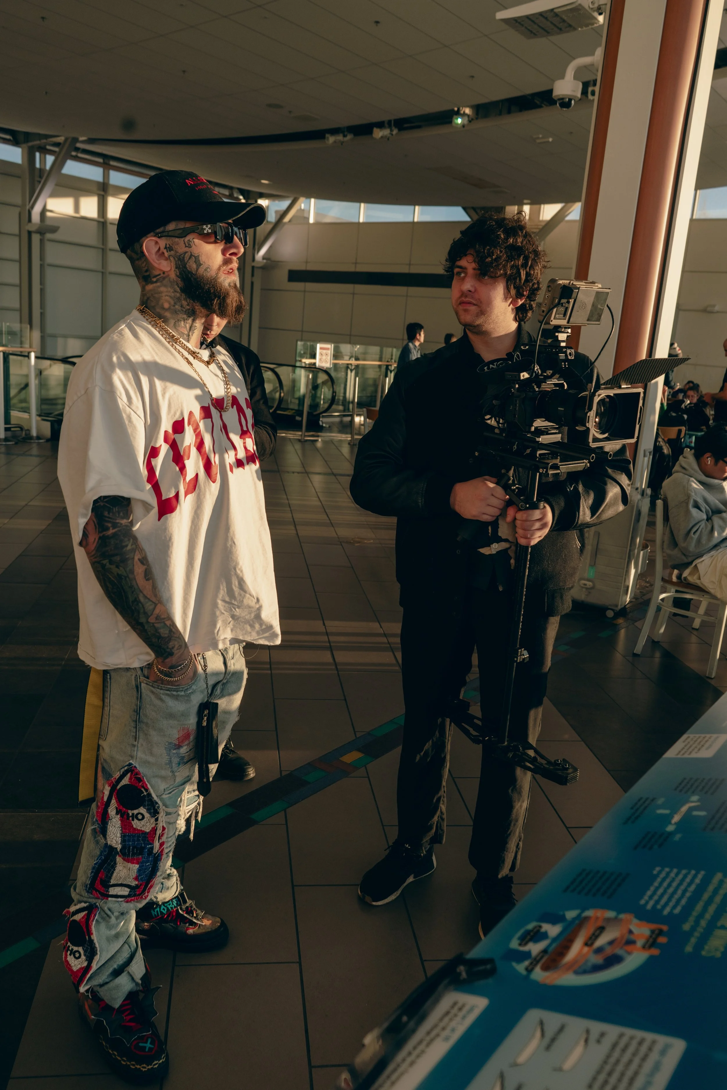 A man with tattoos, wearing sunglasses, a black cap, and a white T-shirt with red lettering, is being filmed by a person operating a professional camera in an indoor public space, possibly an airport.