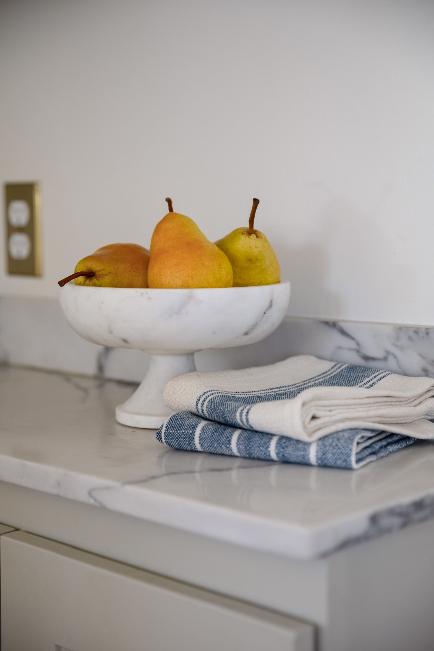 Marble pedestal bowl with pears on quartz countertop beside blue and white dish towels.