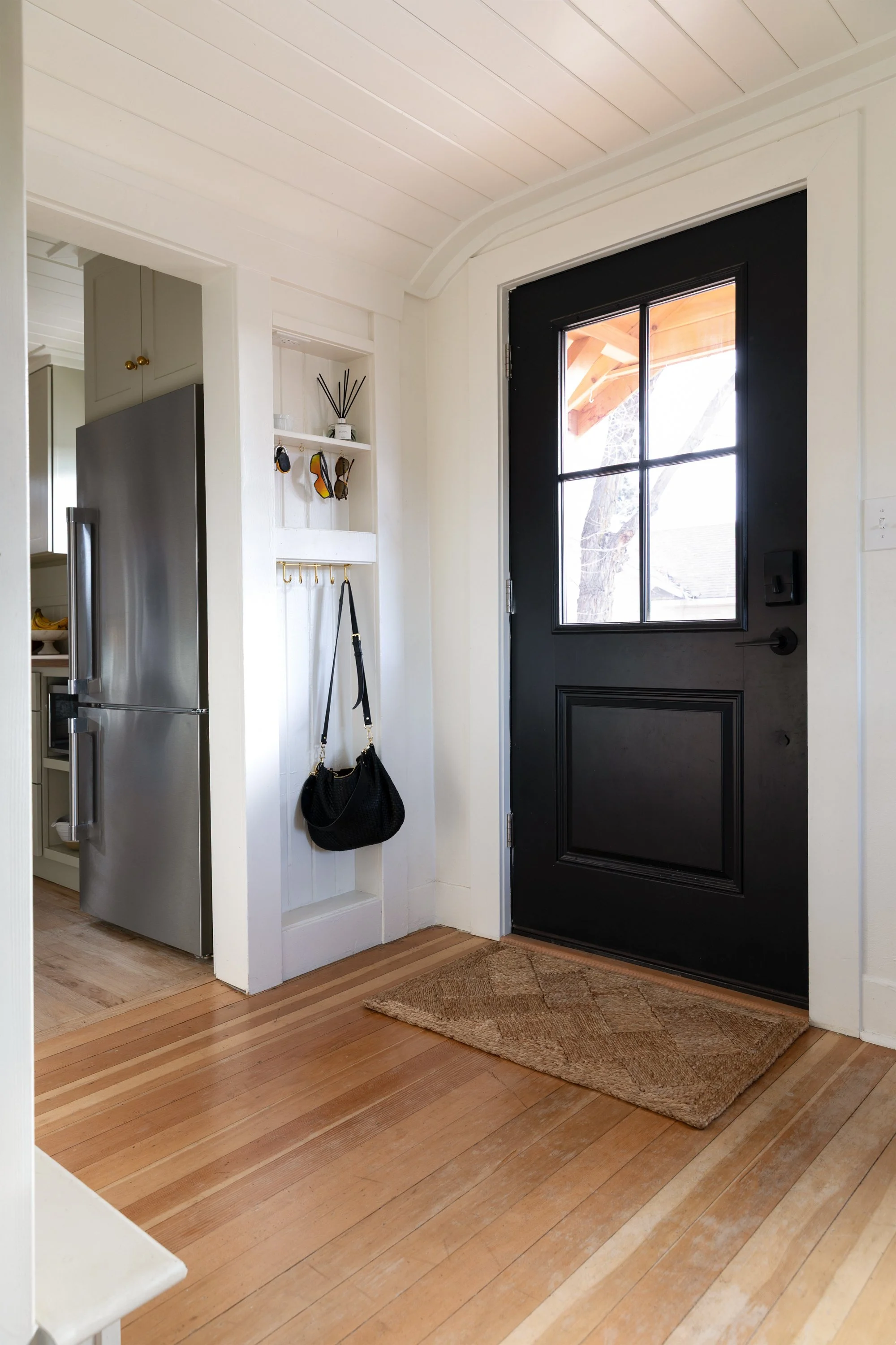 Angled view of the Poplar Cottage front door and small storage built into 2x4 wall with entry rug, black front door, and fir wood floors.