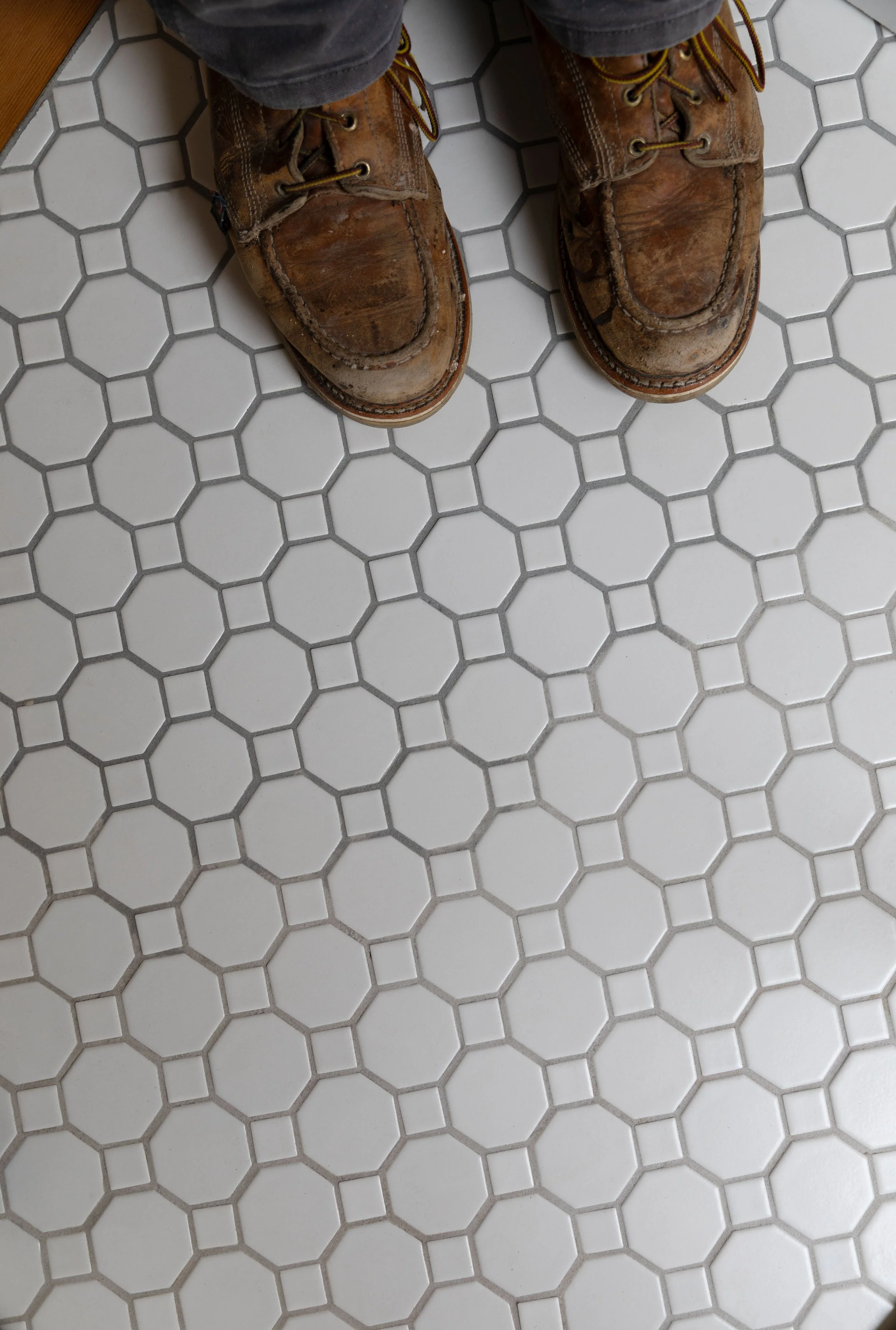 Close-up of bathroom floor tile showing epoxy grout color variation between batches, photographed with boots in frame
