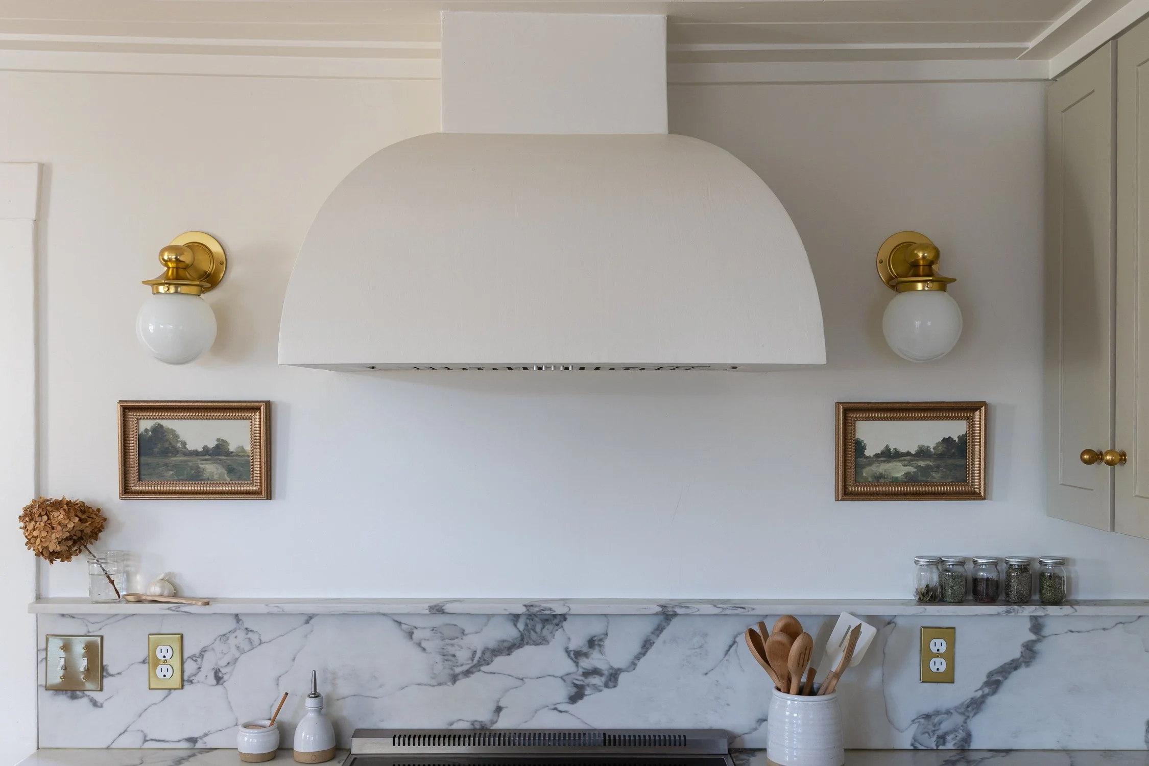 Wide shot of curved plaster range hood with sconces, quartz backsplash, and landscape artwork.