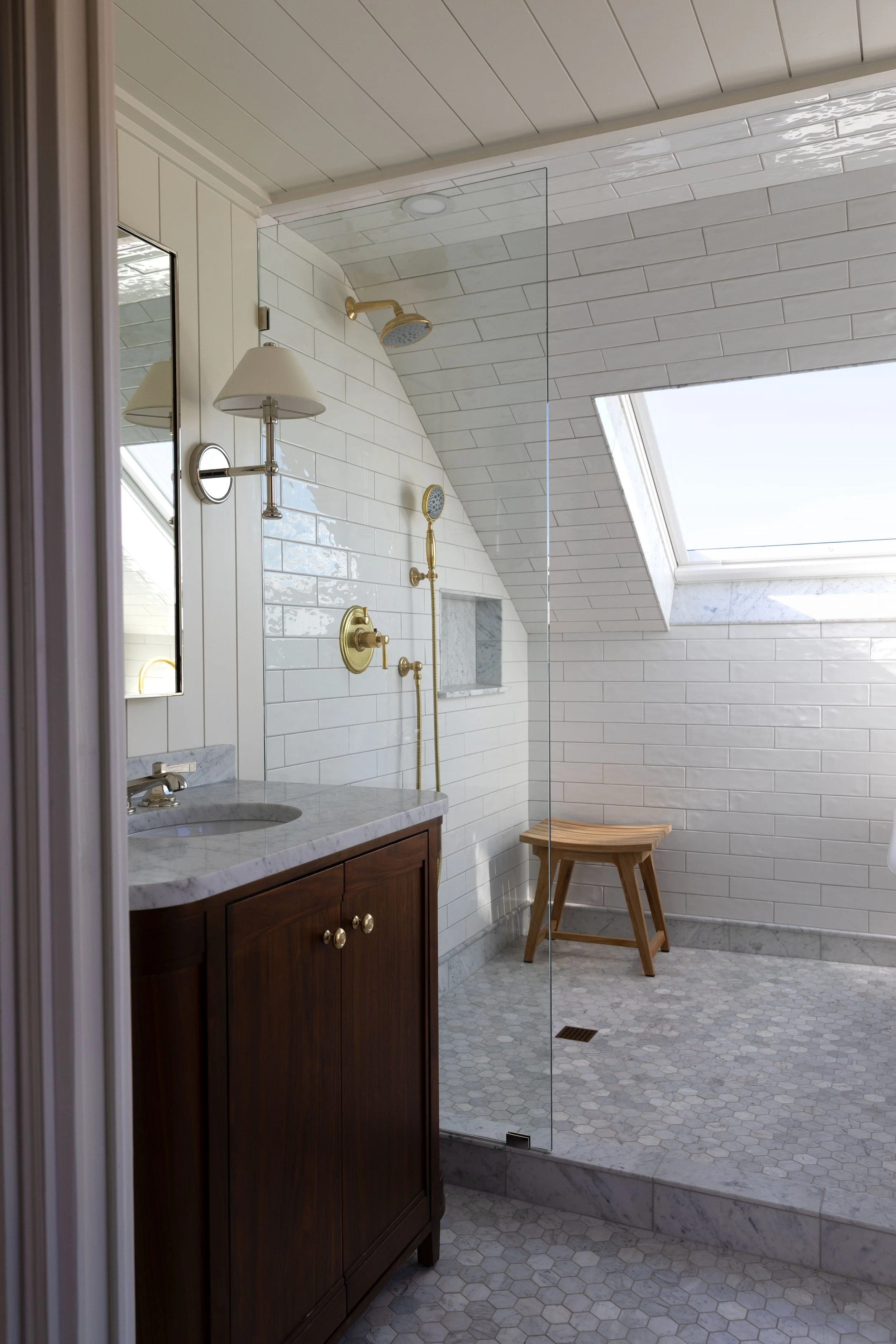 Frameless glass shower panel with brass fixtures in a white bathroom with a skylight