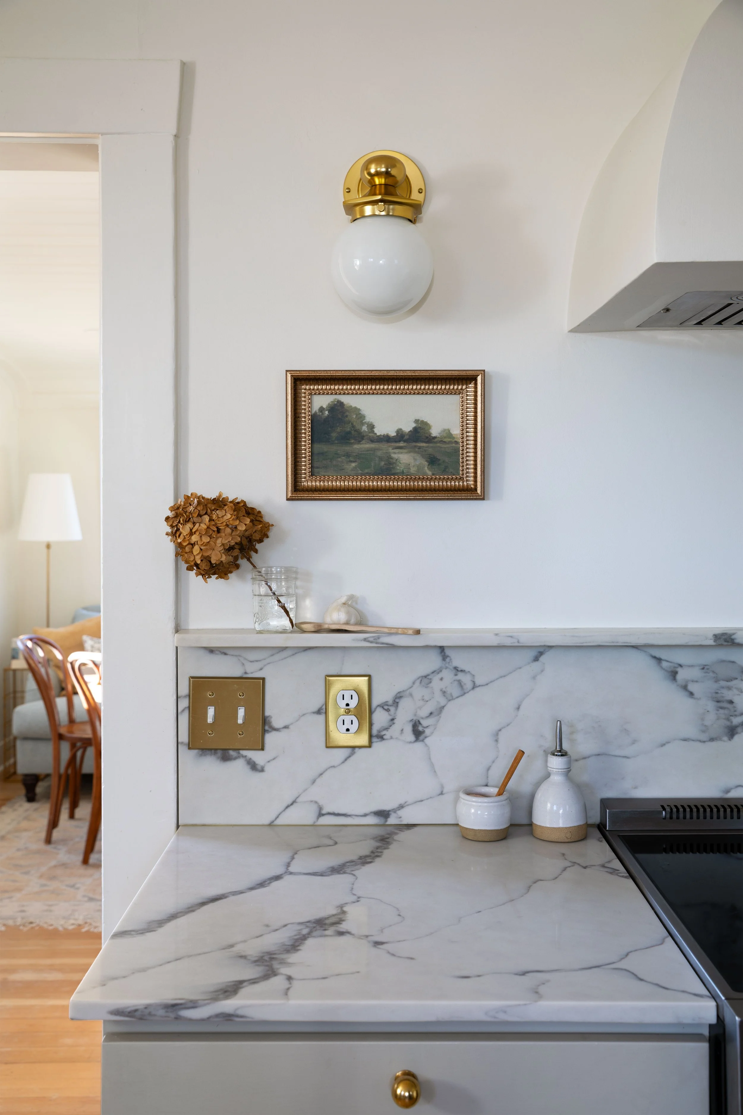 Close-up of quartz backsplash shelf, brass sconce, hydrangea in glass jar, and vintage kitchen artwork.
