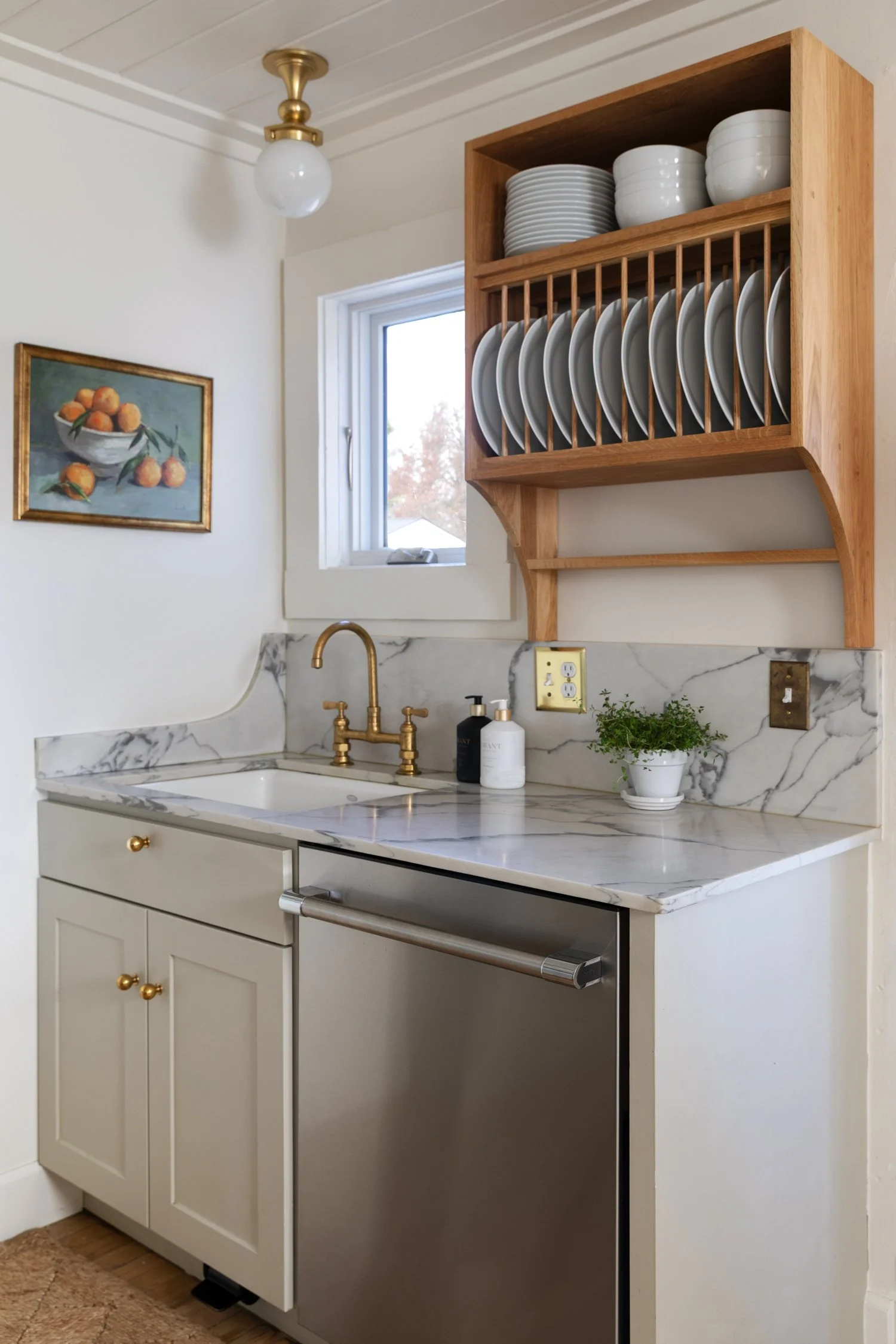 Sink wall with brass faucet, wood plate rack, and dishwasher in cottage kitchen.