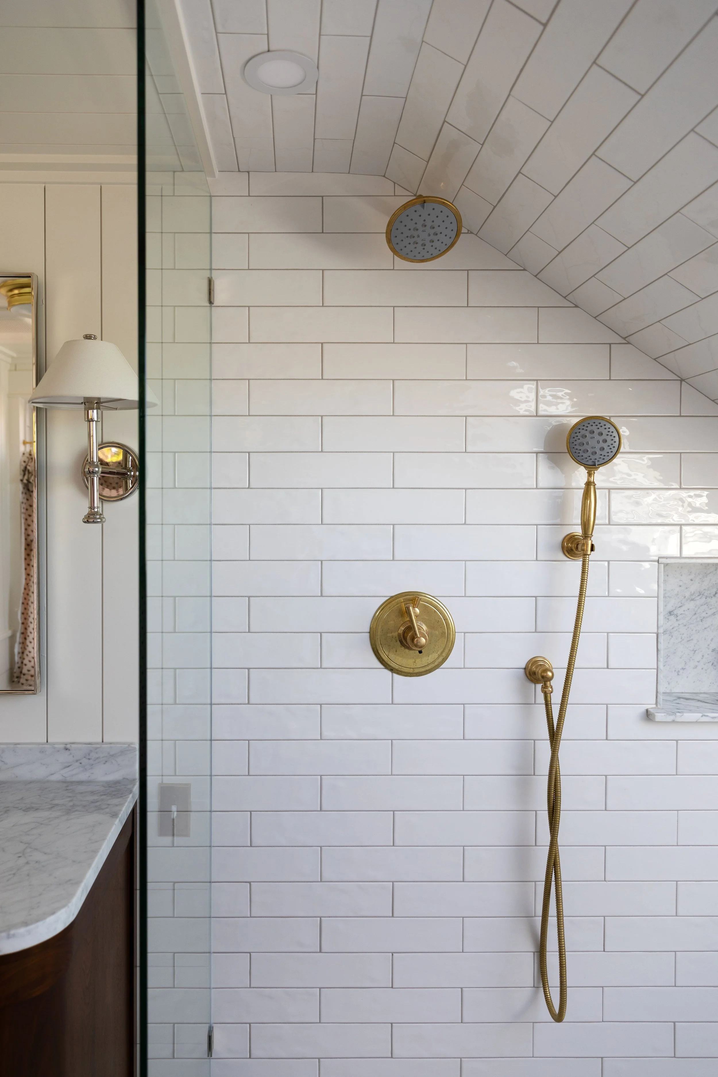 Brass rain shower head and handheld shower against white tile next to a glass panel
