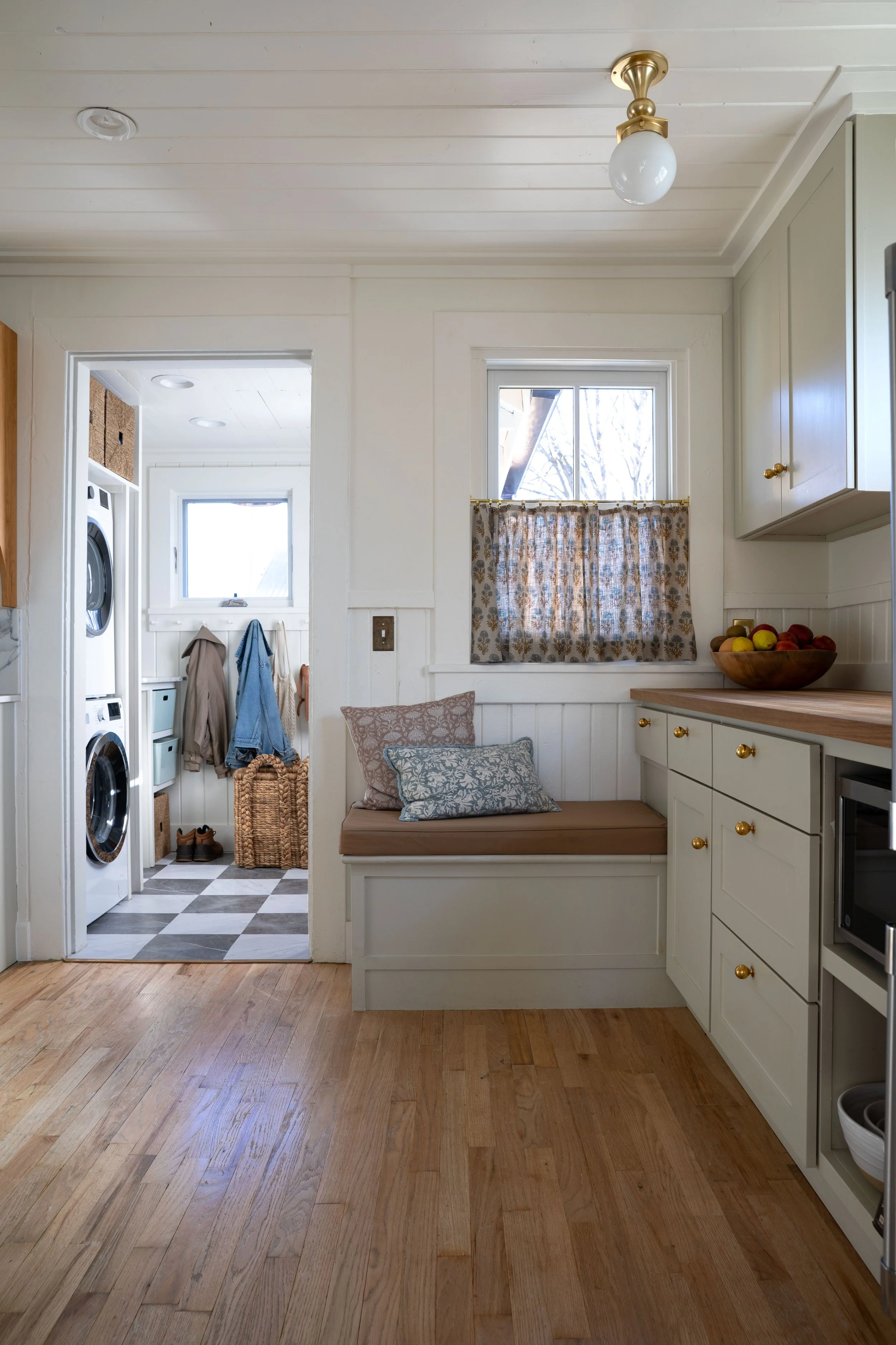 Kitchen view toward mudroom showing built-in bench with cushion and linen café curtains.