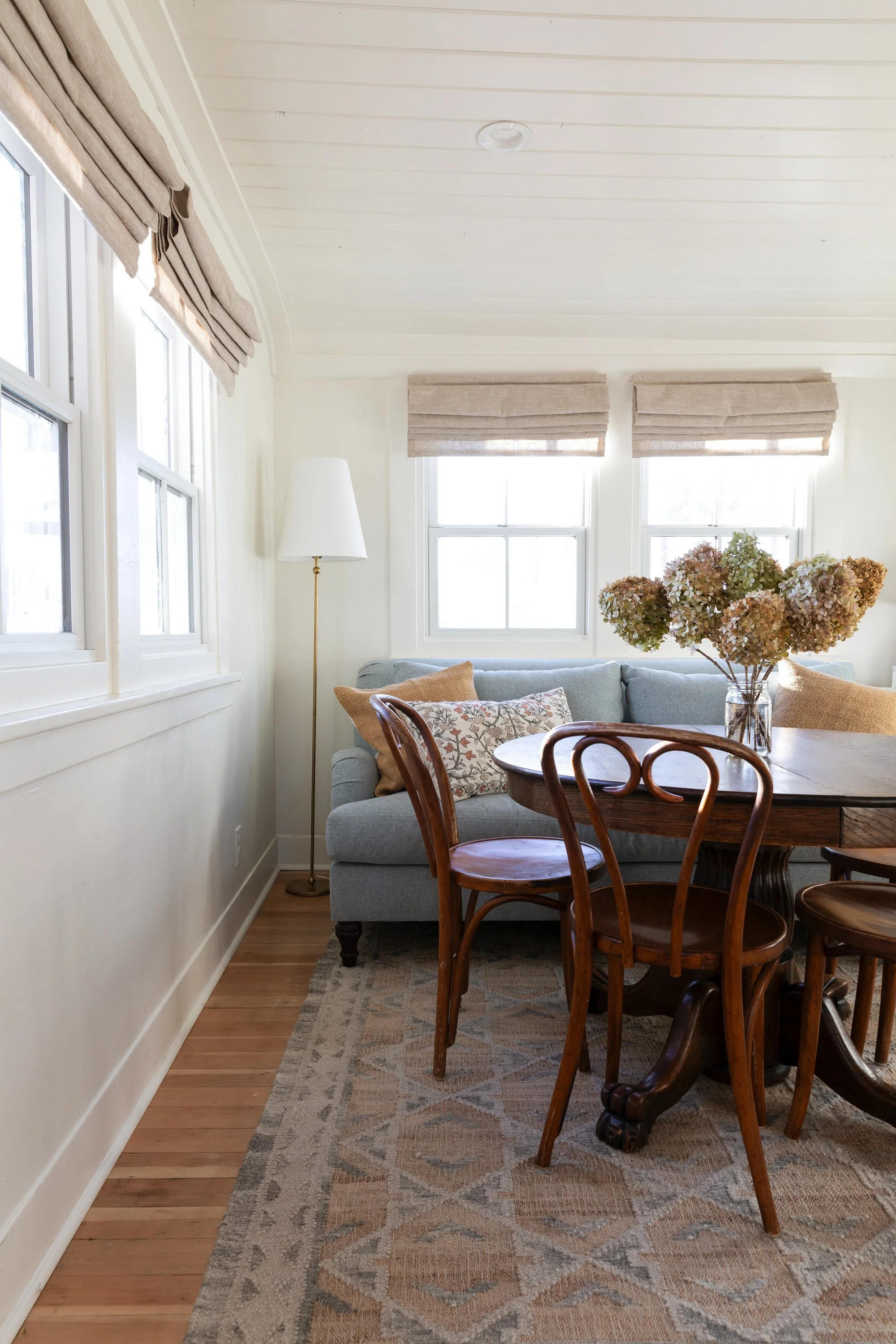 Side view of sitting and dining room with wood-clad windows, bentwood chairs, floor lamp, and blue sofa