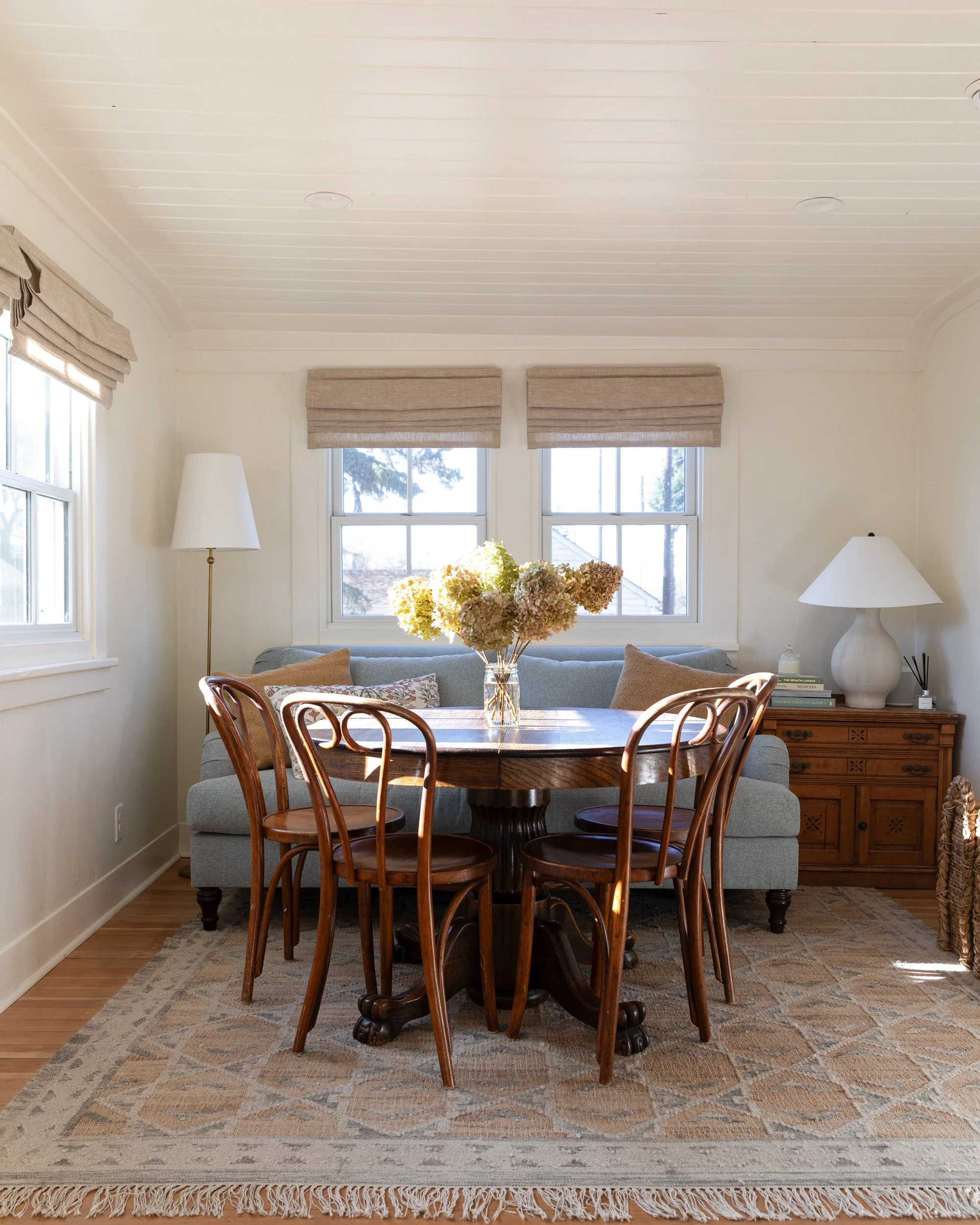 Finished sitting room and dining room combo at the Poplar Cottage with blue sofa, vintage round table, bentwood chairs, and paneled ceiling