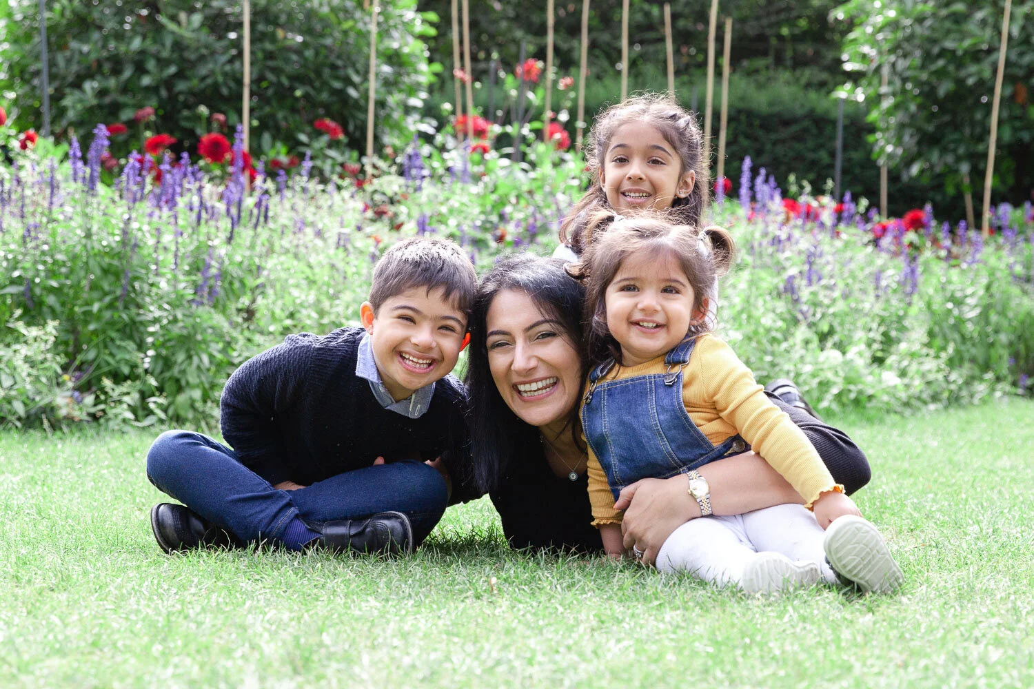 natural family photoshoot in the park with a mum lying down surrounded by her three young children all smiling and looking relaxed.