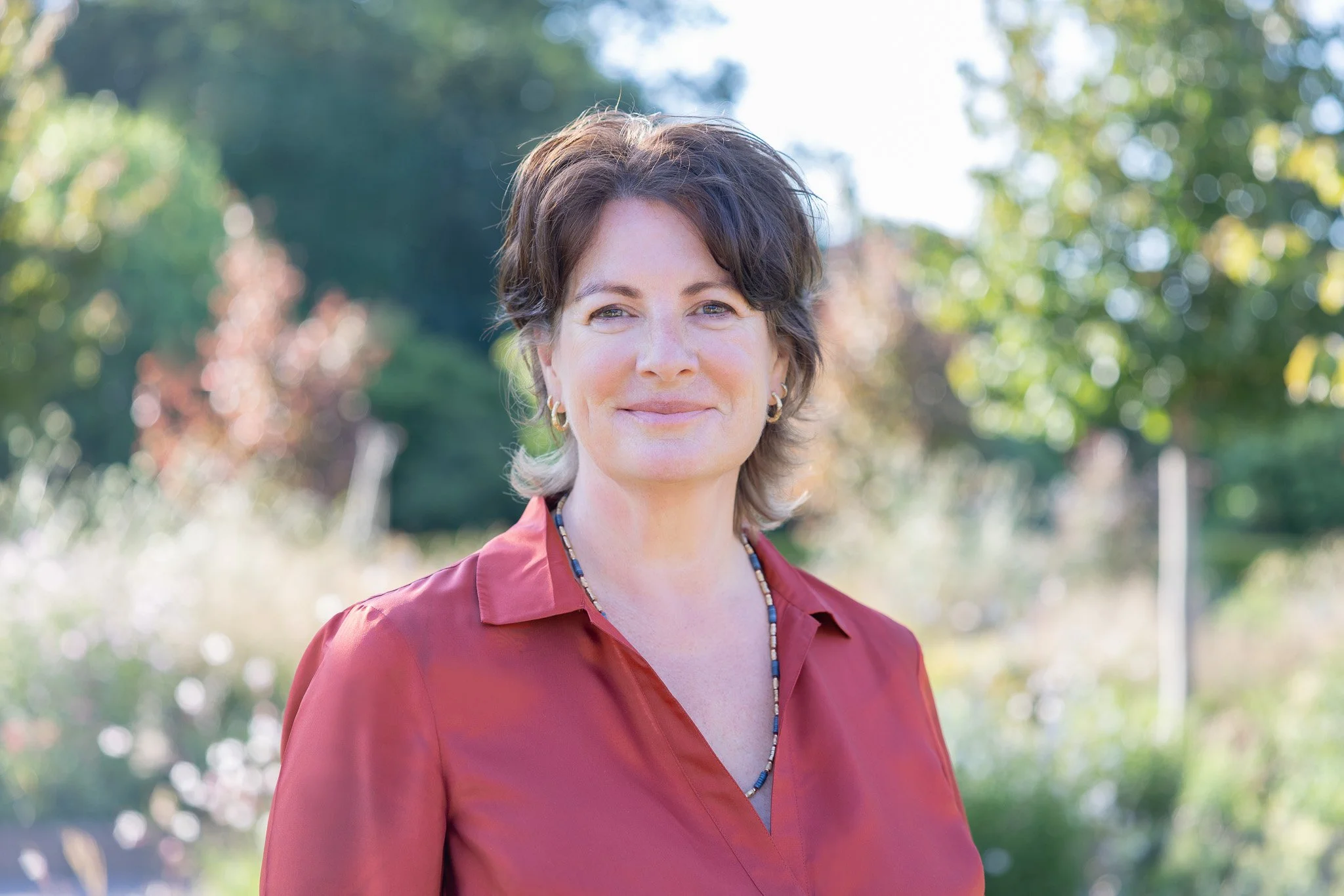 Headshot photography showing a lady wearing a rust coloured shirt. Standing outdoors with greenery behind her. her expression is confident and warm.