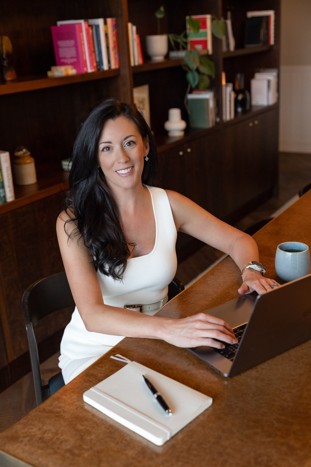 Personal branding photography showing a female business owner sitting at a desk with her computer open. There is a bookshelf behind her and a pen and notebook and coffee cup on the desk. She is looking to the camera with a smiling expression