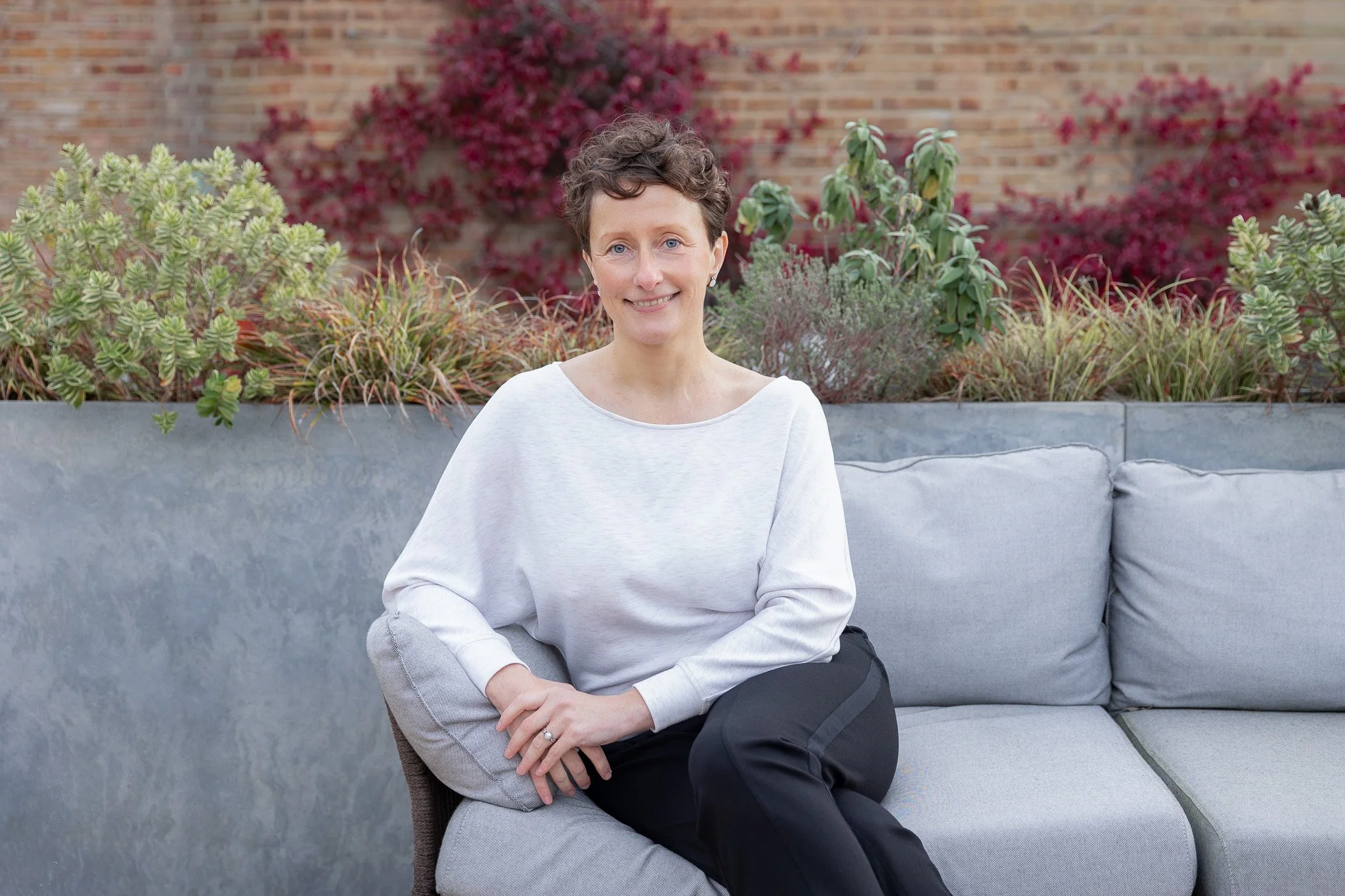 Personal branding photography showing a female business owner sitting on a grey sofa in an outdoor setting. There are planters behind her with plants and a brick wall.