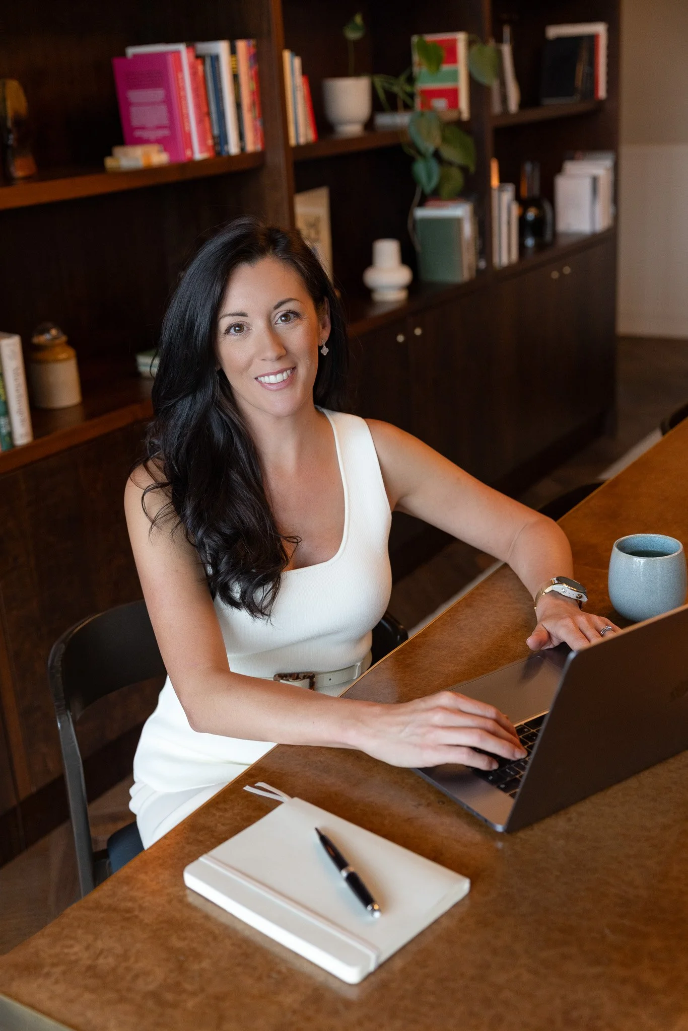 Personal branding photography showing a female business owner sitting at a desk with her computer open. There is a bookshelf behind her and a pen and notebook and coffee cup on the desk. She is looking to the camera with a smiling expression